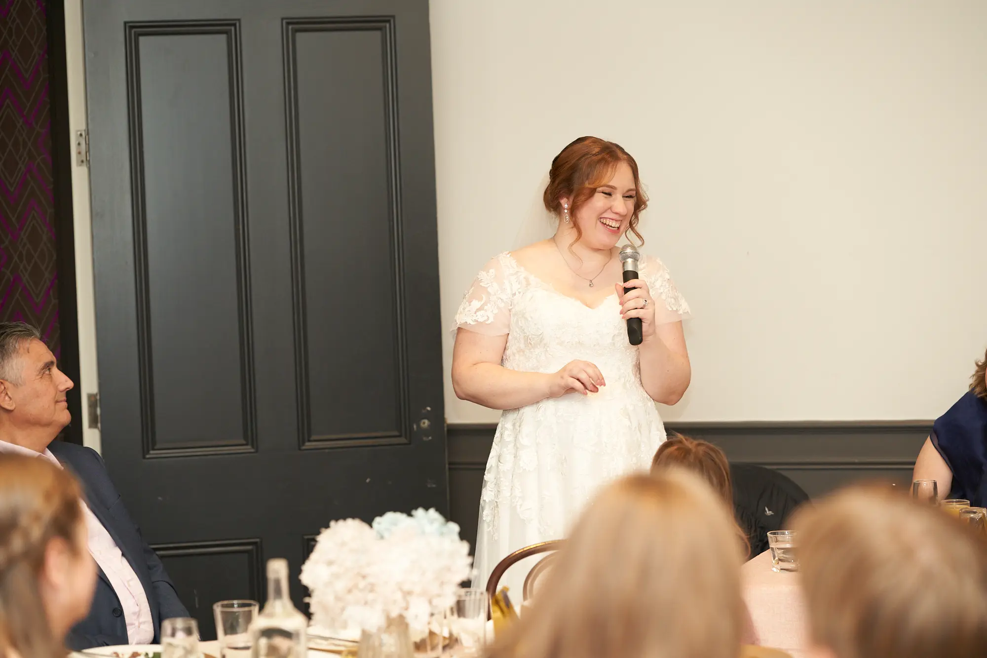 the vincent albert park bride giving speech reception joyful moment A bride gives a heartfelt and joyful speech at her wedding reception at The Vincent, Albert Park. She stands holding a microphone, smiling and engaging her guests. Seated attendees, including a man in a navy suit and a woman in a dark blue dress, listen attentively. The head table features pale pink linens, gold-accented plates, and white and blue floral arrangements. The indoor space has a plain white wall, a black wooden door, and modern purple geometric wallpaper, creating a stylish and intimate setting filled with warmth and celebration.