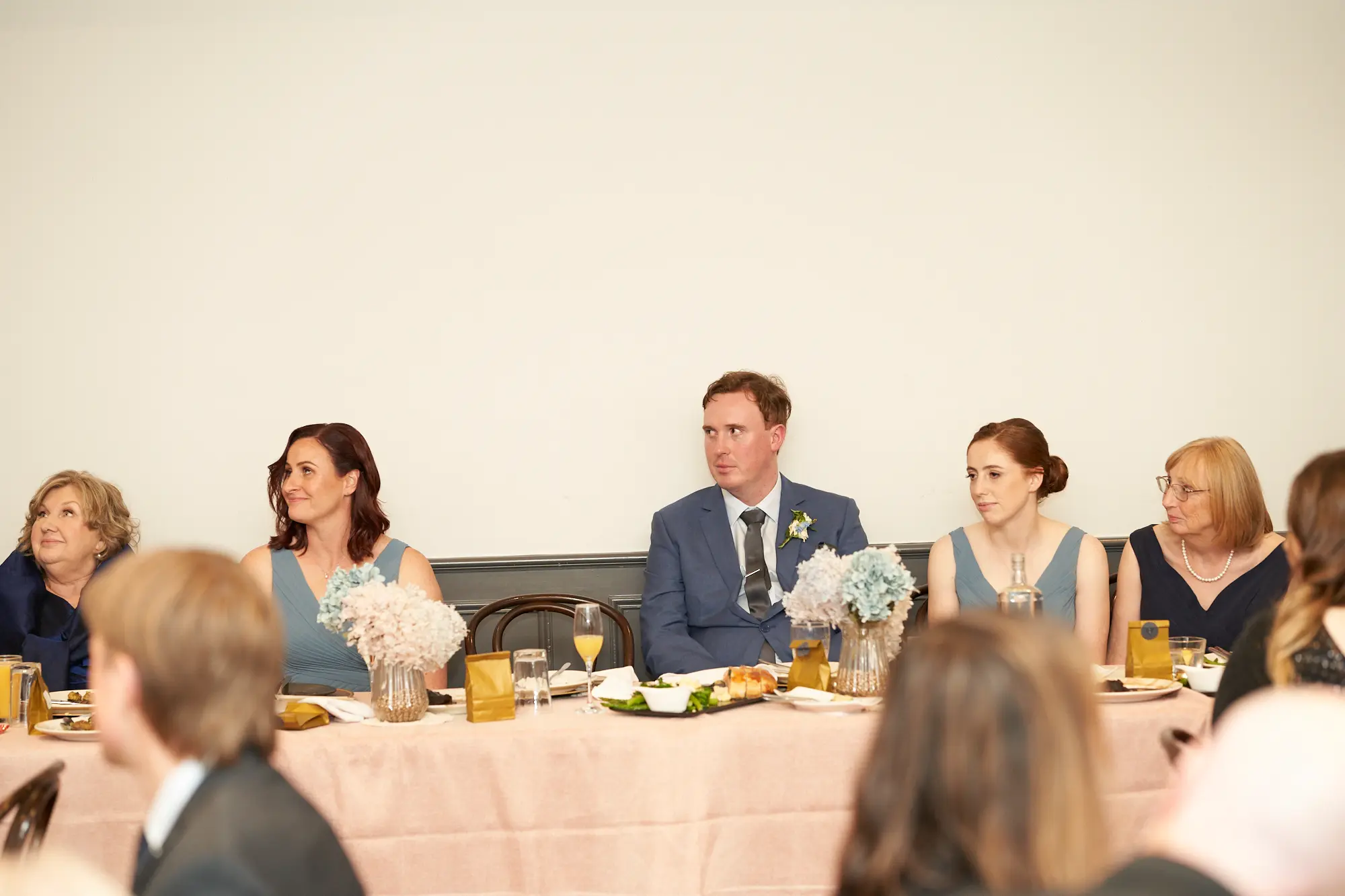 the vincent albert park groom seated head table candid moment The groom sits at the head table during his wedding reception at The Vincent, Albert Park, wearing a light blue suit and patterned tie. Seated guests around him, dressed in dusty blue and navy attire, appear attentive and relaxed. The table is covered with a pale pink cloth, set with gold-accented plates, glassware, and white and blue floral arrangements. The indoor space features a plain white wall and high ceilings, creating an elegant and intimate atmosphere that captures a candid moment of calm enjoyment and shared happiness during the celebration.