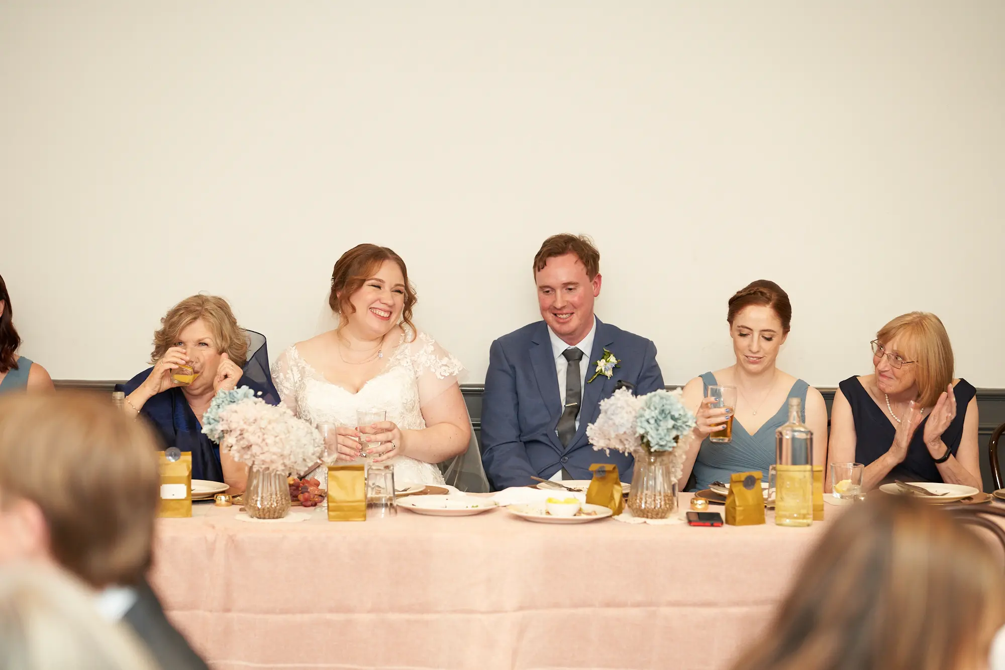 the vincent albert park wedding reception head table joyful laughter floral elegance A joyful candid moment at The Vincent in Albert Park showing the newly married couple laughing at the head table, surrounded by smiling guests. Five people sit along a long table decorated with pale pink linens, gold-accented plates, and pastel floral arrangements. The bride laughs openly while the groom smiles warmly forward. Guests on either side look toward the couple with affection. The bright white wall, high ceilings, and soft lighting create an elegant, intimate atmosphere during the wedding reception.