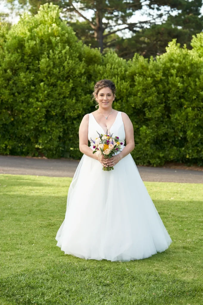 The bride poses elegantly for a portrait on the manicured lawn of Farm Vigano, wearing a white gown with a deep V-neckline and holding a vibrant, multi-colored bouquet. She smiles warmly at the camera, standing confidently in front of lush green hedges and mature trees. Bright natural sunlight illuminates the scene under a clear blue sky, creating a joyful, serene, and romantic atmosphere that captures her happiness and grace on her wedding day.