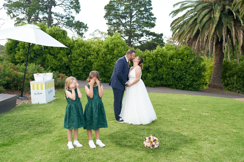 svg%3E Allison and Christian share a tender kiss after their outdoor wedding ceremony at Farm Vigano in South Morang, Melbourne, while two young flower girls in matching dark green dresses playfully cover their eyes, with one peeking through her fingers. Set on a manicured lawn surrounded by lush trees, a vibrant palm, and scenic views of the Plenty Valley Gorge, the moment blends romance, humor, and family warmth, creating a lighthearted, intimate, and timeless celebration under bright daylight.