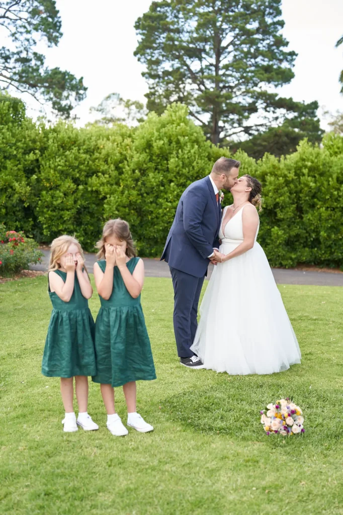 The bride and groom share a tender, celebratory kiss on the manicured lawn of Farm Vigano, the bride holding a light-colored bouquet resting on the grass. In the foreground, two young flower girls in dark green dresses cover their eyes, one peeking through her fingers, adding a playful, adorable touch. Behind them, lush hedges, mature trees, a vibrant palm, and a white sun umbrella with a yellow cart create a scenic, sun-drenched setting. The bright daylight and natural surroundings capture a joyful, intimate, and lighthearted moment full of romance and family charm.