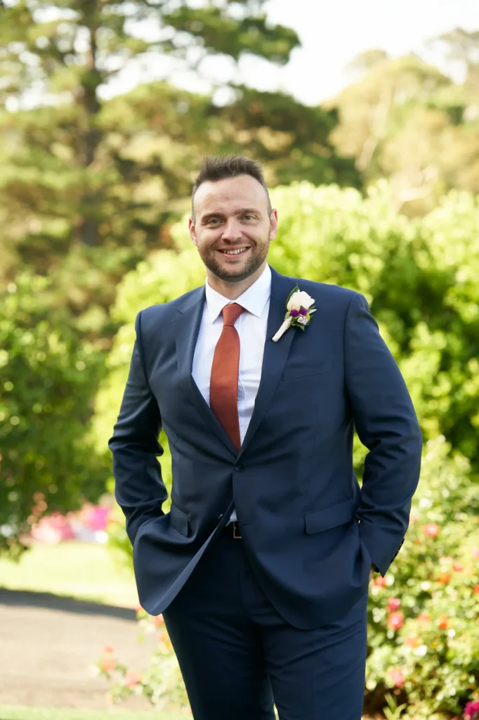 The groom poses confidently for a portrait at Farm Vigano, standing on a manicured lawn surrounded by lush green hedges and mature trees. He wears a sharp navy suit, white shirt, and rust-colored tie, with a boutonniere of purple and white flowers on his lapel. Both hands are casually tucked into his pockets as he smiles warmly at the camera. Hints of colorful garden flowers in the foreground add vibrancy, while bright natural daylight illuminates the scene, creating an elegant, joyful, and relaxed atmosphere in this Italian-inspired outdoor setting.