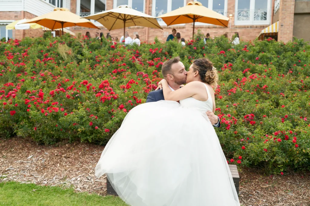 svg%3E Allison and Christian share a joyful, passionate kiss as the groom lifts the bride in his arms in front of a vibrant bed of red roses at Farm Vigano, South Morang, Melbourne. Her white dress billows elegantly as guests watch from a patio with yellow umbrellas, surrounded by lush greenery and Italian-inspired architecture, capturing a playful, romantic, and intimate celebration bathed in natural sunlight.