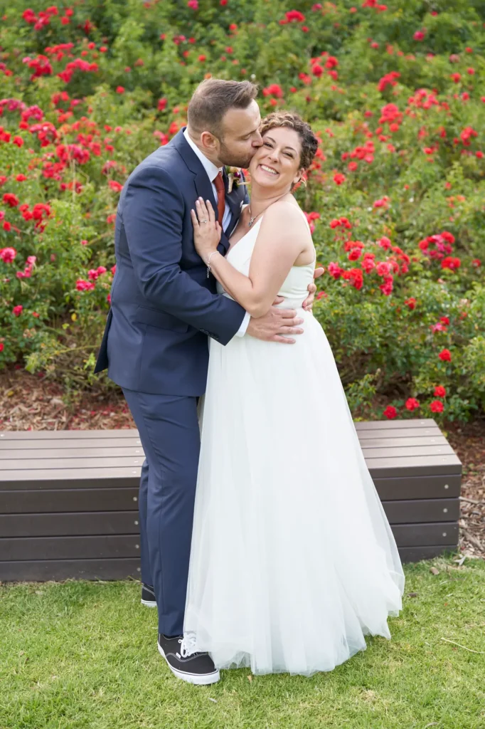 The bride and groom stand closely on the manicured lawn of Farm Vigano, the groom tenderly kissing the bride on the cheek while she smiles with pure happiness. He wears a navy suit with a rust-colored tie and black sneakers, and she wears a white gown with a deep V-neckline, her arm around his waist. Behind them, a dense wall of vibrant red roses and lush greenery forms a striking backdrop, with a dark brown garden bench in the mid-ground, capturing a joyful, intimate, and romantic moment in the elegant outdoor setting of this Italian-inspired wedding.