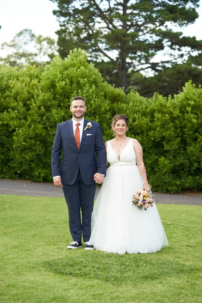 The bride and groom stand side-by-side on the manicured lawn of Farm Vigano, holding hands and smiling warmly at the camera. The groom wears a sharp navy suit with a rust-colored tie and black sneakers, while the bride wears a white gown with a deep V-neckline and a full skirt, holding a colorful bouquet. Behind them, a dense wall of lush green hedges and mature trees provides a natural, elegant backdrop, with a large tree in the upper background. The bright sunlight and open-air setting create a joyful, classic, and romantic wedding portrait.