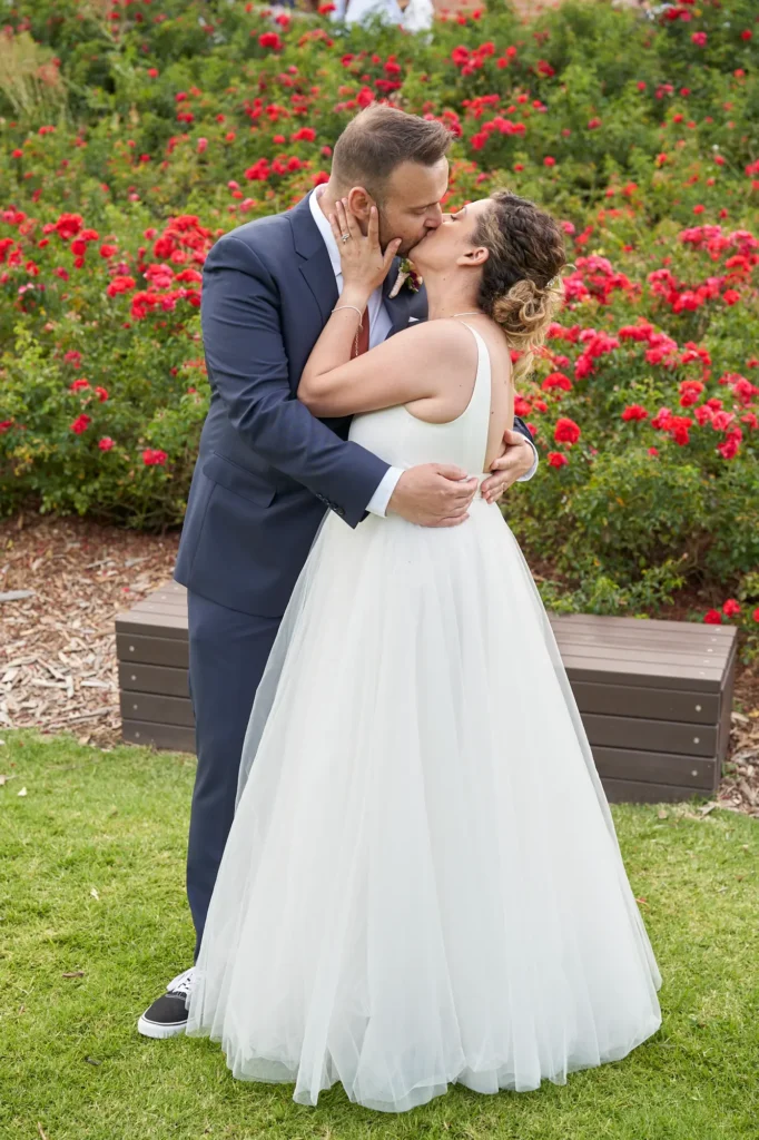 The bride and groom share a tender, celebratory kiss on a manicured lawn at Farm Vigano. The groom wears a navy suit with a rust-colored tie and black sneakers, holding the bride close by her waist. The bride, in a white gown with a deep V-neckline and full skirt, has her right arm around his neck and holds a vibrant bouquet in her left hand. Behind them, a dark brown garden bench sits in front of a dense wall of red roses and lush green foliage, with the building and guests faintly visible in the sunlit background, creating a romantic and intimate atmosphere.