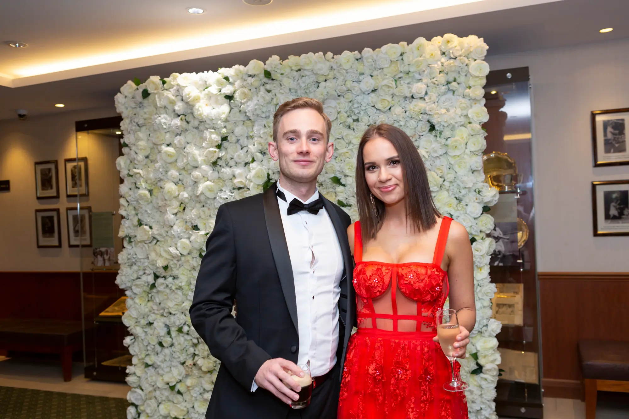kooyong lawn tennis club end of year masquerade ball couple red dress A couple poses together at the Kooyong Lawn Tennis Club End-of-Year Masquerade Ball in front of a white and cream rose floral wall. The man wears a classic black tuxedo with a bow tie, and the woman wears a red strappy gown with a sheer lace bodice, each holding a glass. Smiling confidently at the camera, they capture the festive, sophisticated, and celebratory atmosphere of this prestigious Melbourne end-of-year masquerade event.