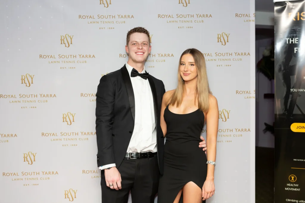 svg%3E A stylish couple pose together in front of a white and cream rose floral wall with Royal South Yarra Lawn Tennis Club banners during a formal end-of-year celebration. Dressed in classic black-tie attire, the man in a tuxedo and the woman in an elegant black strapless gown smile confidently at the camera, reflecting the refined, social atmosphere of a prestigious Melbourne tennis club event that celebrates connection, tradition, and elegant evening entertainment.