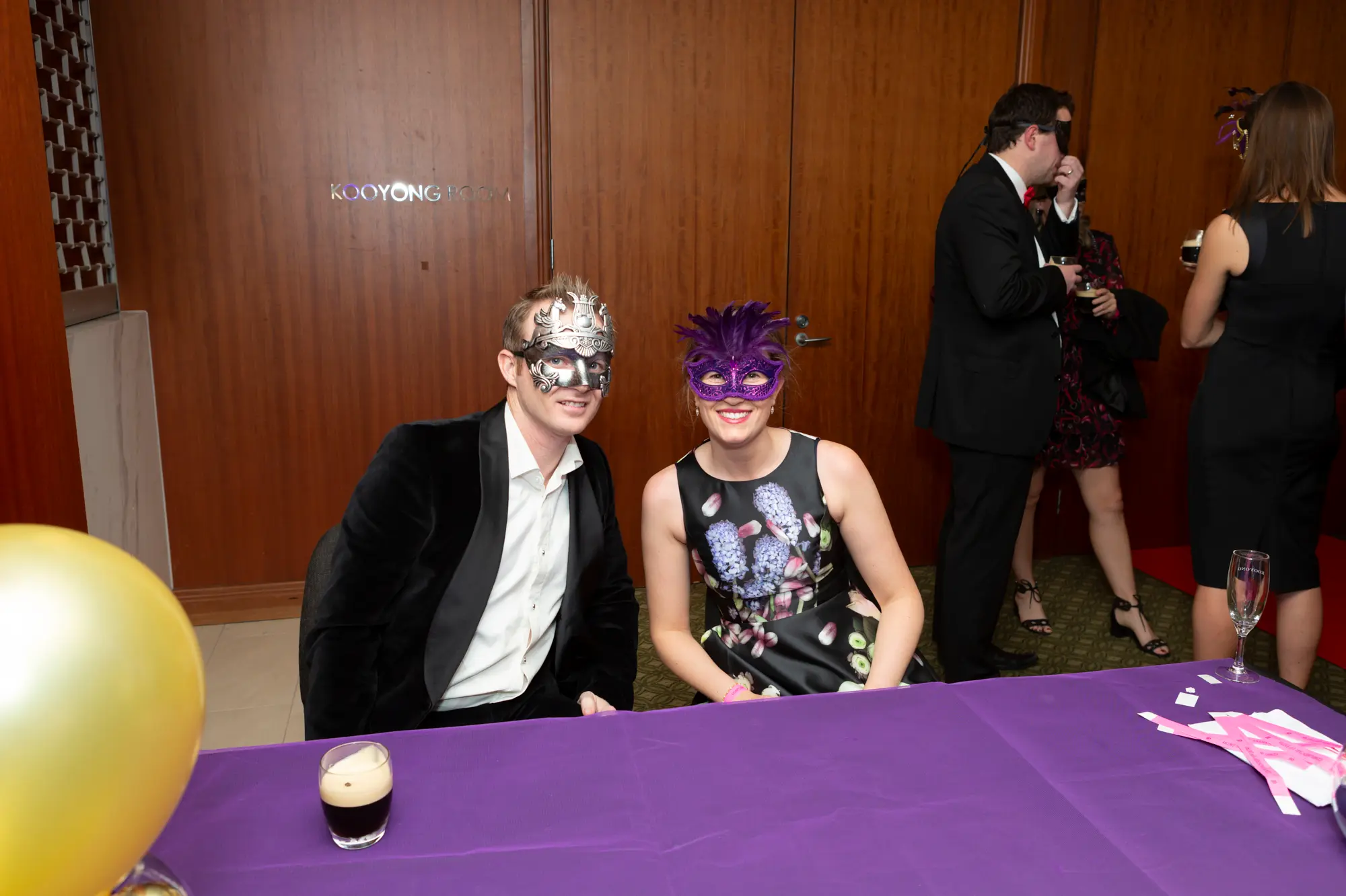 kooyong lawn tennis club end of year masquerade ball couple seated purple table A couple is seated at a purple-covered table at the Kooyong Lawn Tennis Club End-of-Year Masquerade Ball, smiling at the camera. The man wears a black velvet jacket, white shirt, and ornate silver masquerade eye mask, while the woman wears a sleeveless black floral dress with a purple feathered mask. Surrounded by other formally dressed guests, drinks on the table, and wood-paneled walls, they convey a festive, sophisticated, and celebratory atmosphere at this prestigious Melbourne end-of-year masquerade event.