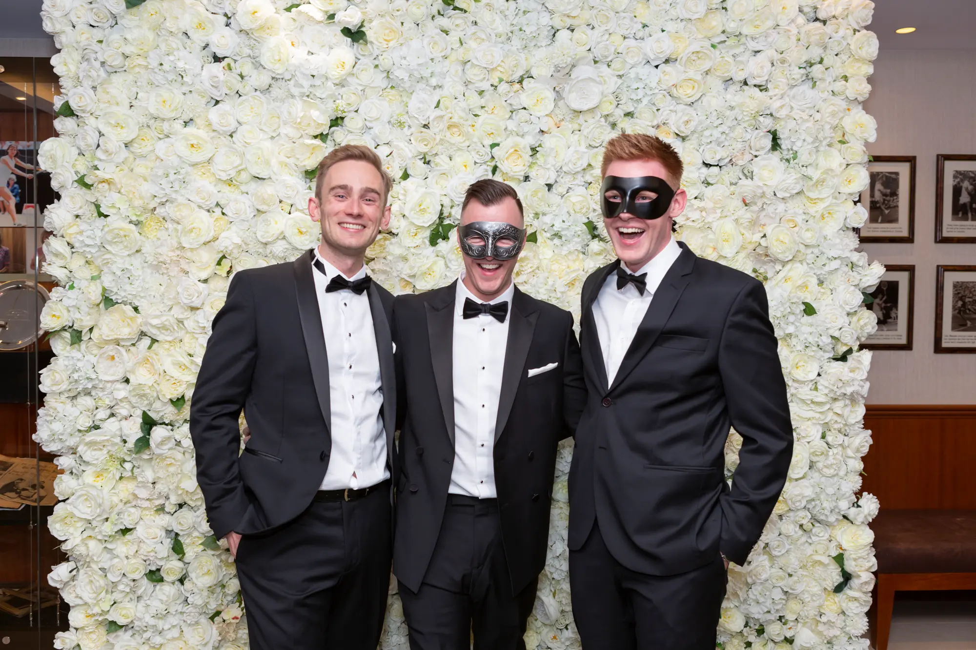kooyong lawn tennis club end of year masquerade ball elegant arrivals three men smile Three men pose together at the Kooyong Lawn Tennis Club End-of-Year Masquerade Ball in front of a white and cream rose floral wall with wood paneling and framed pictures. Dressed in black tuxedos with bow ties and masquerade eye masks, the man in the center wearing a waistcoat, they smile confidently at the camera, capturing the sophisticated, festive, and glamorous atmosphere of this prestigious Melbourne end-of-year masquerade event.