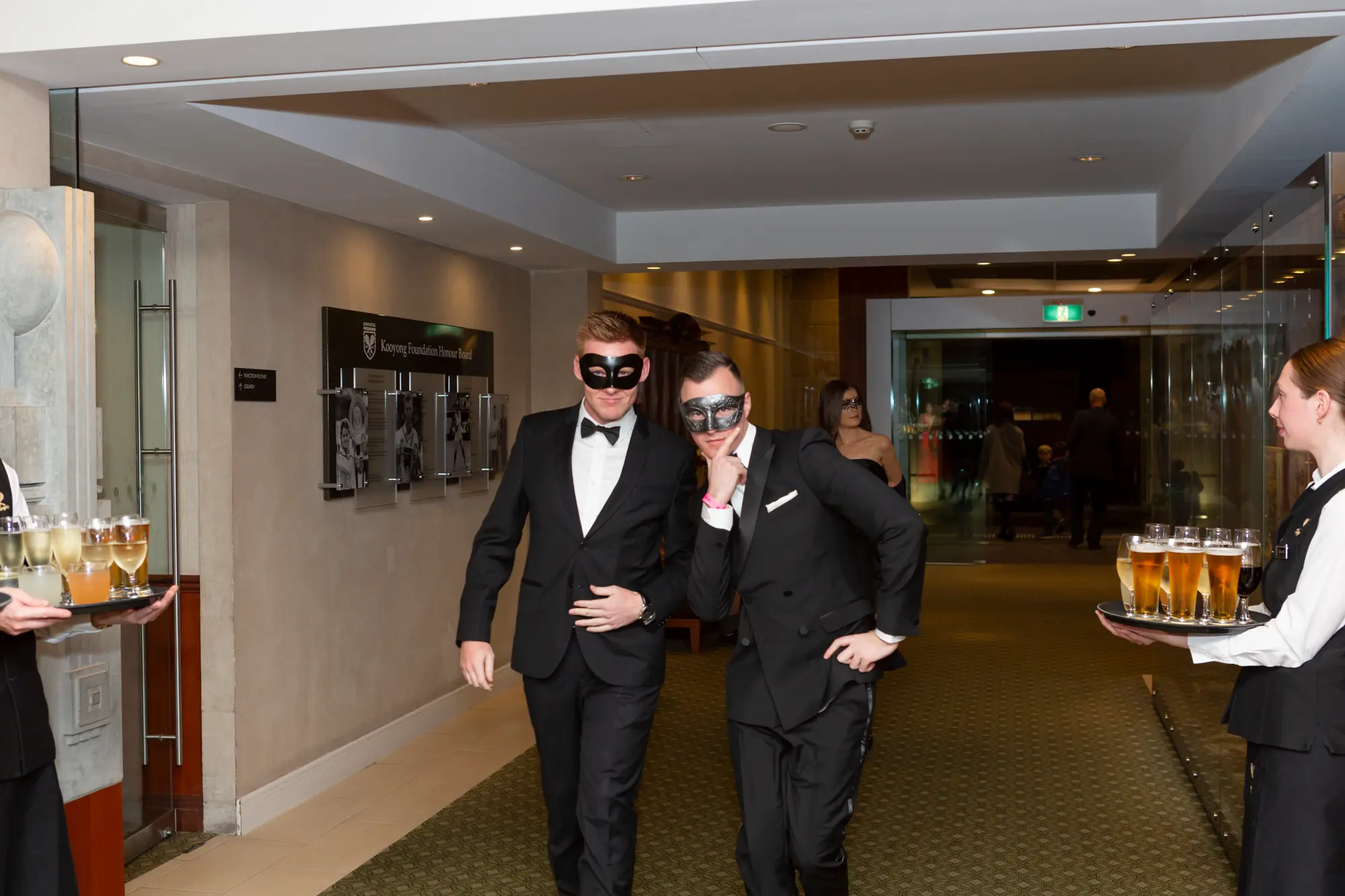 kooyong lawn tennis club end of year masquerade ball elegant arrivals two men entrance Two men make an elegant entrance at the Kooyong Lawn Tennis Club End-of-Year Masquerade Ball, pausing on a red carpet in front of the club’s interior and a glimpse of a white and cream rose floral wall. Dressed in black tuxedos with bow ties and masquerade eye masks, they smile at the camera, capturing the festive, sophisticated, and celebratory atmosphere of this prestigious Melbourne end-of-year masquerade event, with professional attendants ready to serve drinks nearby.