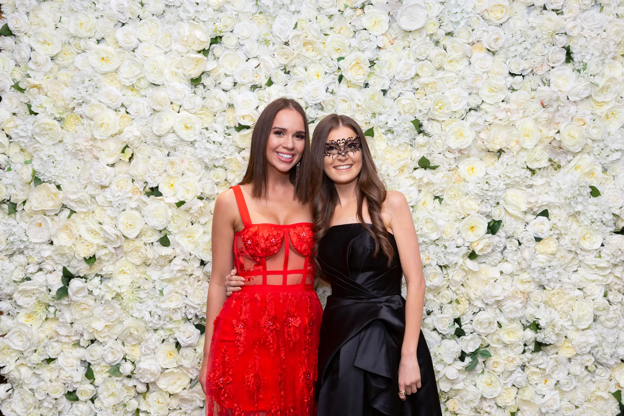 kooyong lawn tennis club end of year masquerade ball elegant arrivals two women navy Two women pose together at the Kooyong Lawn Tennis Club End-of-Year Masquerade Ball in front of a white and cream rose floral wall. Dressed in formal evening wear with masquerade eye masks, one in a long-sleeved navy blue sequined gown and the other in a light patterned strapless dress, they link arms and smile confidently at the camera, capturing the festive, sophisticated, and glamorous atmosphere of this prestigious Melbourne end-of-year masquerade event.