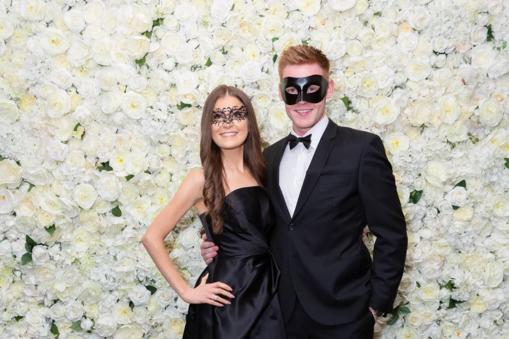 svg%3E A well-dressed couple pose confidently in front of a white and cream rose floral wall at the Kooyong Lawn Tennis Club End-of-Year Masquerade Ball in Toorak. Dressed in elegant black formalwear and wearing classic black masquerade masks, they smile warmly at the camera, capturing the refined and celebratory atmosphere of one of Melbourne’s most prestigious private club events, where sophistication, connection, and timeless social tradition come together.