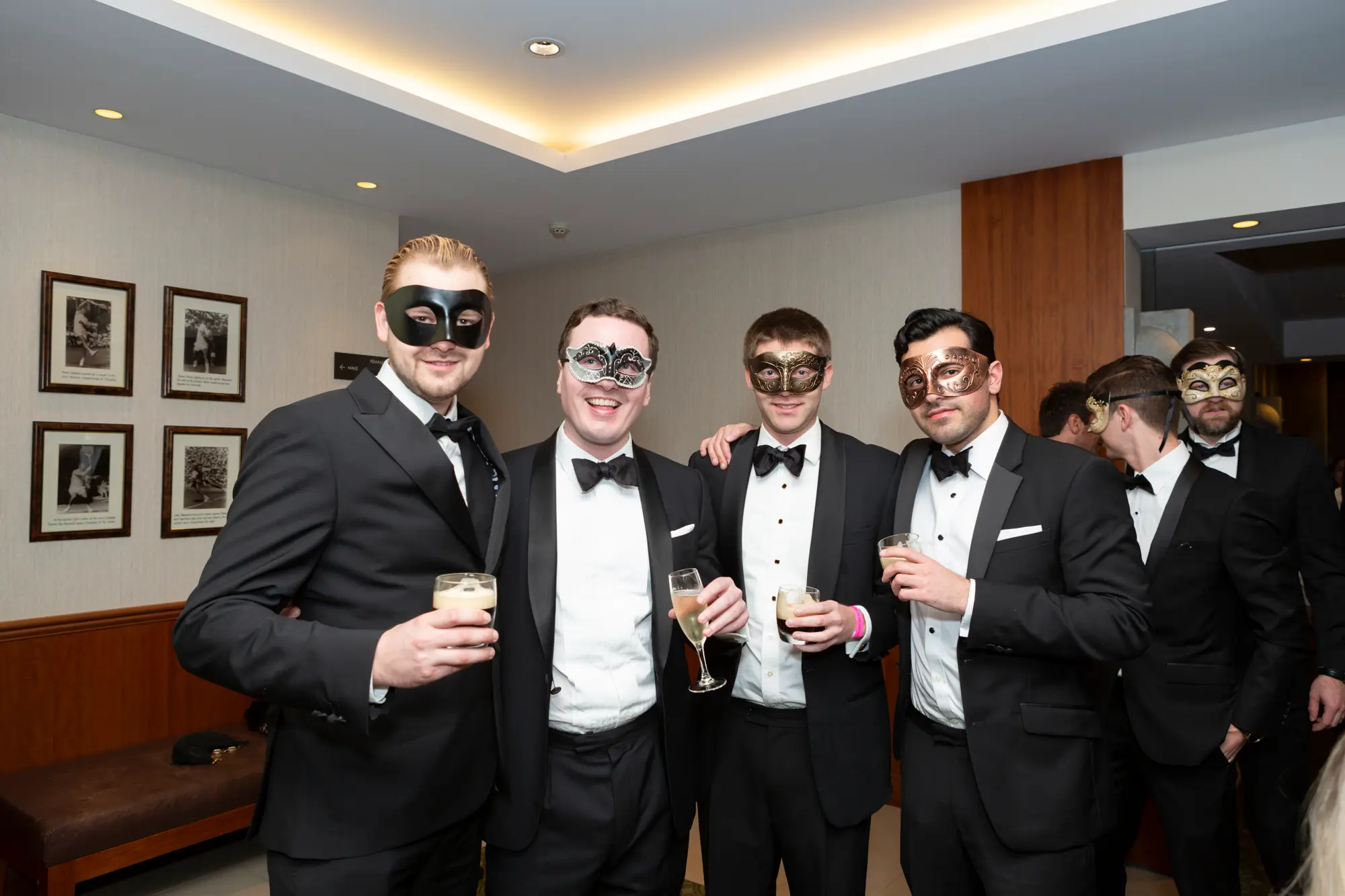kooyong lawn tennis club end of year masquerade ball four men entrance Four men pose together at the Kooyong Lawn Tennis Club End-of-Year Masquerade Ball near an entrance with green-patterned carpet and wood-paneled walls. Dressed in black tuxedos with black bow ties and wearing black or ornate masquerade eye masks, they hold glasses with beverages and smile confidently at the camera. The bright, elegant event space in the background, including glimpses of other guests and the rose wall photo backdrop, conveys a festive, sophisticated, and celebratory atmosphere typical of this prestigious Melbourne end-of-year masquerade event.