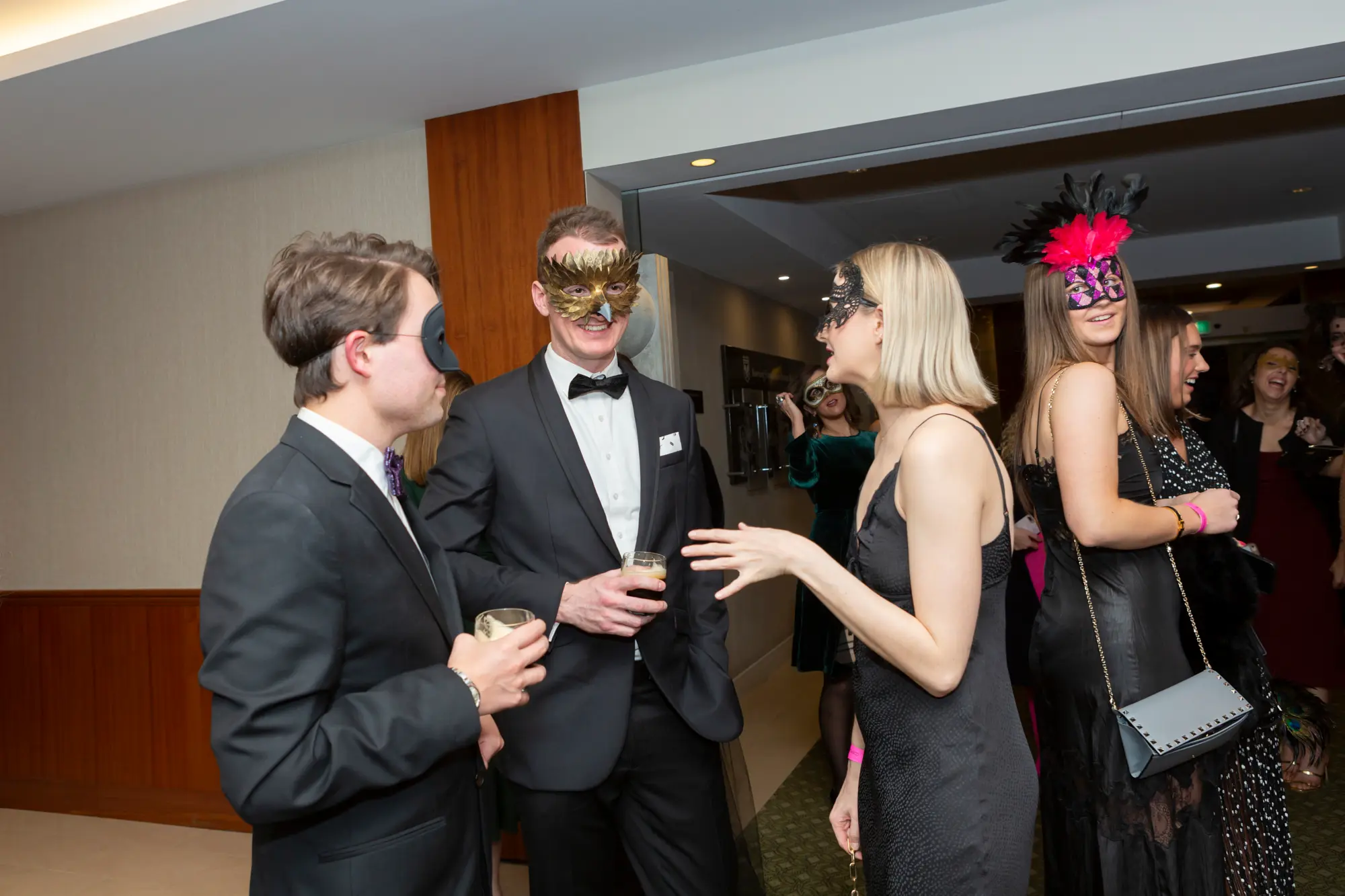 kooyong lawn tennis club end of year masquerade ball group five masquerade A group of five guests poses and converses at the Kooyong Lawn Tennis Club End-of-Year Masquerade Ball in an indoor event space with wood paneling and green-patterned carpet. Dressed in formal evening wear with elaborate masquerade eye masks, the two men wear tuxedos with bow ties, while the three women wear elegant black dresses, one with a pink and black feathered mask. They smile and engage with each other, capturing the festive, sophisticated, and celebratory atmosphere of this prestigious Melbourne end-of-year masquerade event.