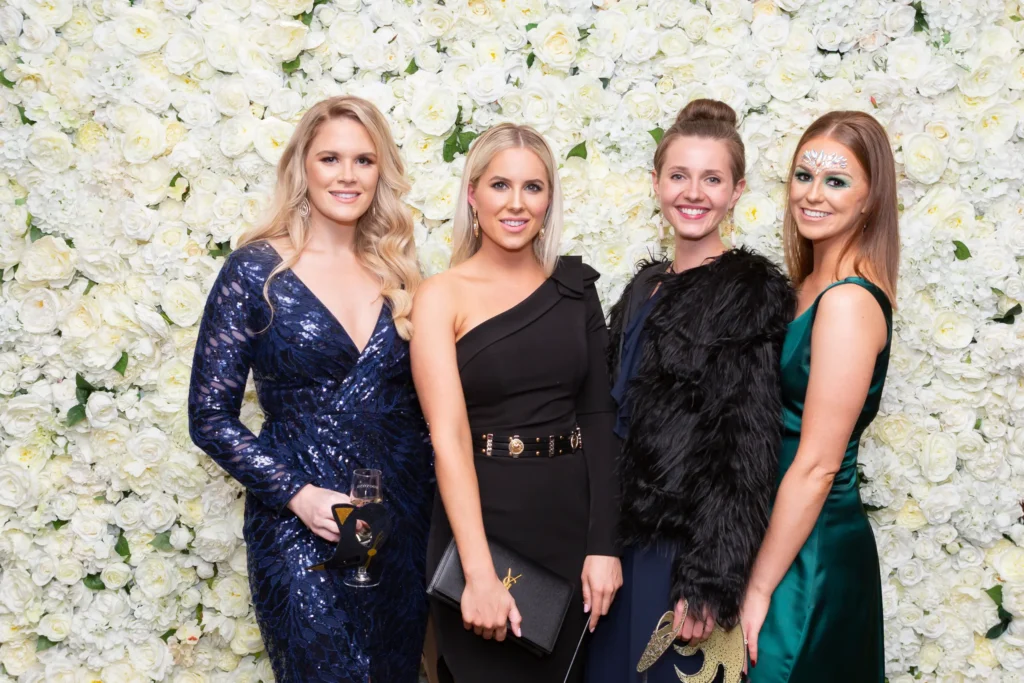 kooyong lawn tennis club end of year masquerade ball group of women floral wall Four elegantly dressed women pose together in front of a white and cream rose floral wall at the Kooyong Lawn Tennis Club End-of-Year Masquerade Ball in Toorak. Wearing formal evening gowns, sequins, masks, and delicate masquerade face paint, they smile warmly at the camera, capturing the glamorous, social, and celebratory spirit of this prestigious end-of-year event filled with style, friendship, and refined Melbourne society.