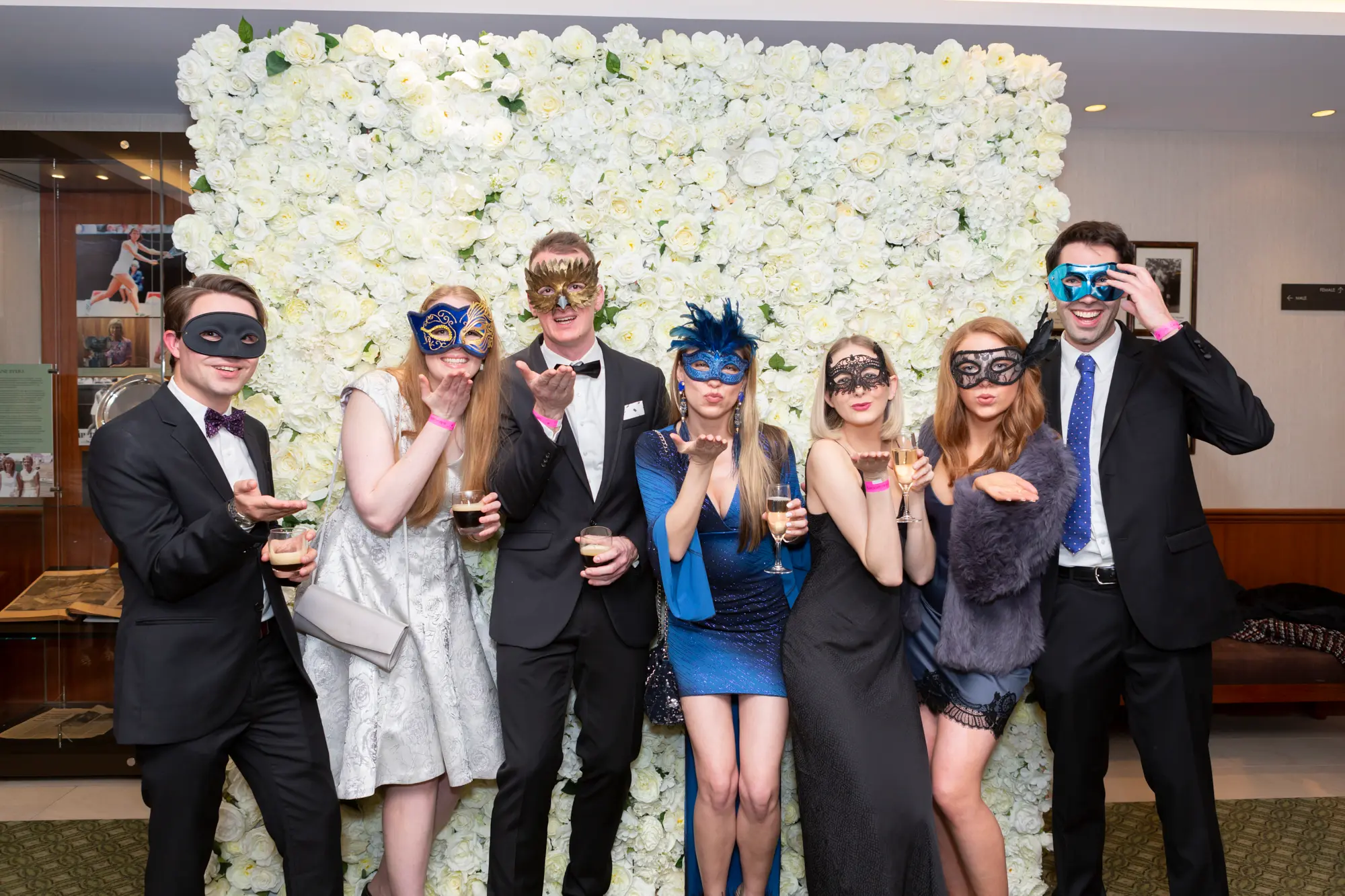 kooyong lawn tennis club end of year masquerade ball group seven rose wall Seven guests pose together at the Kooyong Lawn Tennis Club End-of-Year Masquerade Ball in front of a white and cream rose floral wall with wood paneling and framed pictures. Dressed in formal evening wear with masquerade eye masks, the men wear tuxedos with bow ties, and the women wear elegant dresses, including a dark sequined gown and a blue feathered mask. Smiling confidently at the camera, they capture the festive, sophisticated, and celebratory atmosphere of this prestigious Melbourne end-of-year masquerade event.