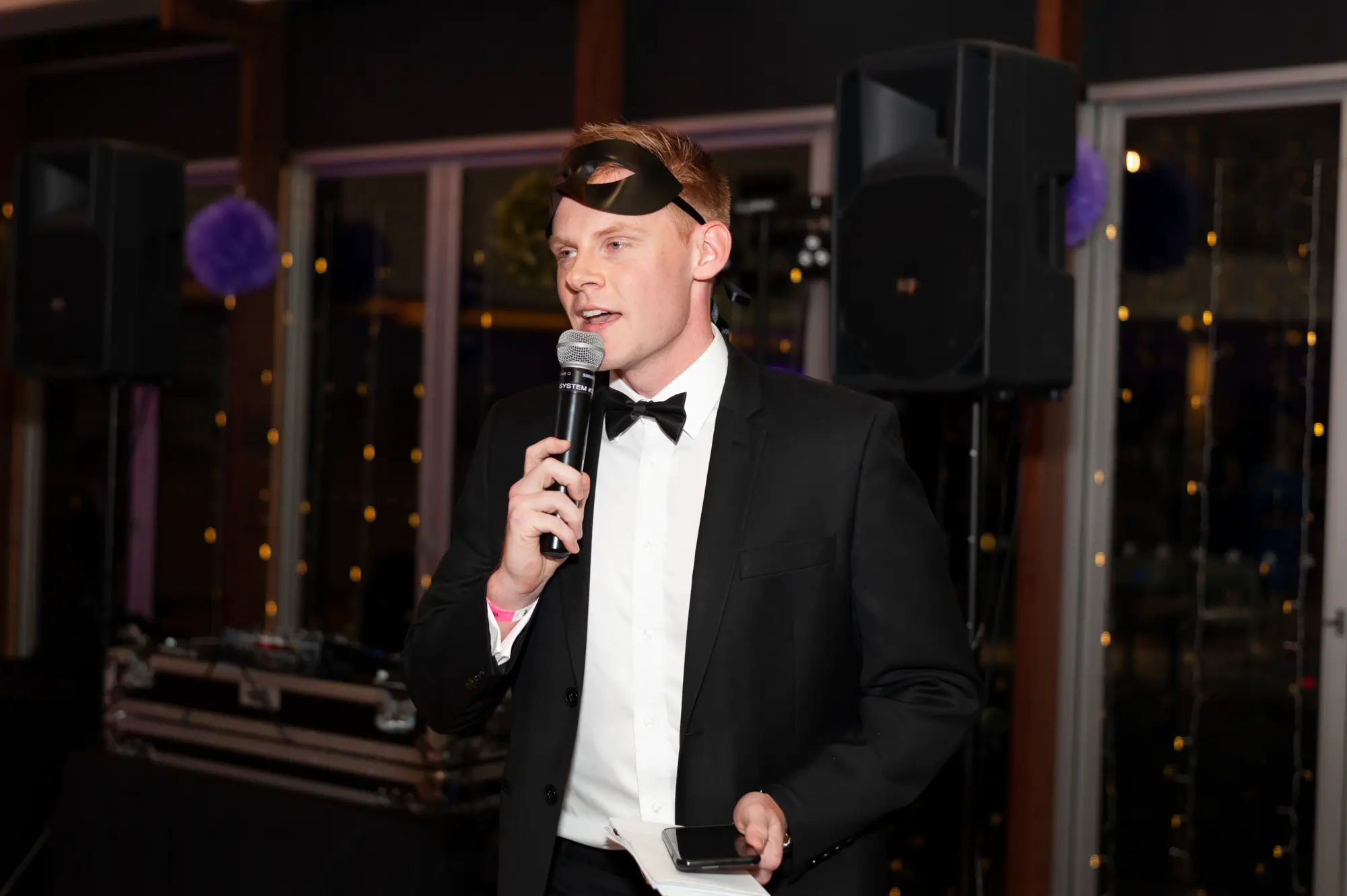 kooyong lawn tennis club end of year masquerade ball speeches main room A masked host in a black tuxedo delivers an engaging speech into a microphone during the Kooyong Lawn Tennis Club End-of-Year Masquerade Ball in Melbourne, standing in the main event room as guests gather for formal speeches, drinks, and shared moments beneath elegant lighting and professional audio equipment.