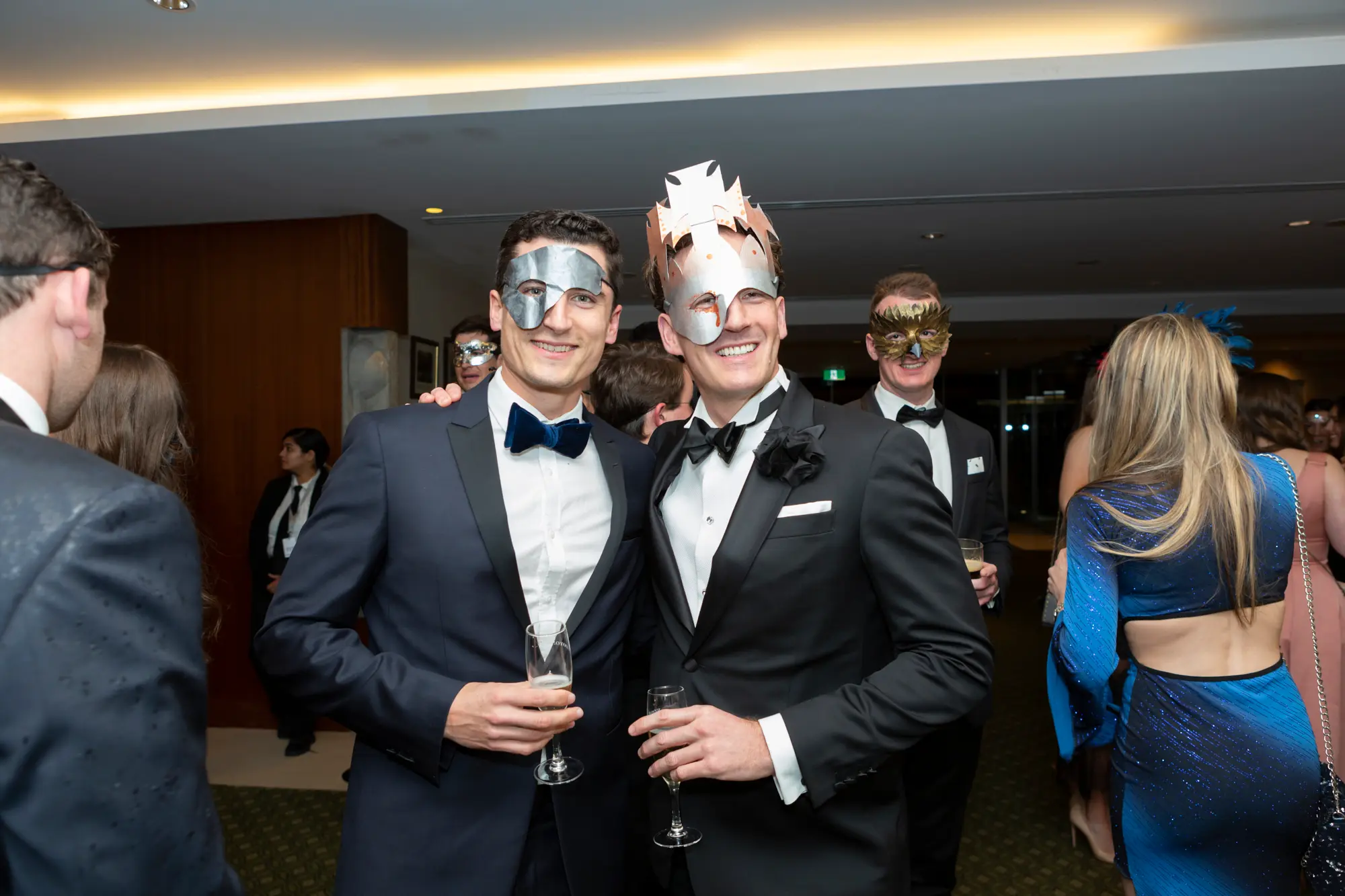 kooyong lawn tennis club end of year masquerade ball two men indoor event wo men pose together at the Kooyong Lawn Tennis Club End-of-Year Masquerade Ball in a sophisticated indoor event space with wood paneling and patterned carpet. Dressed in formal evening wear with ornate masquerade eye masks, one in a navy blue tuxedo with blue bow tie and silver mask holding a glass of champagne, and the other in a black tuxedo with black bow tie and a silver crown-style mask holding a beverage, they smile confidently at the camera, reflecting the festive, social, and celebratory atmosphere of this prestigious Melbourne end-of-year masquerade event.