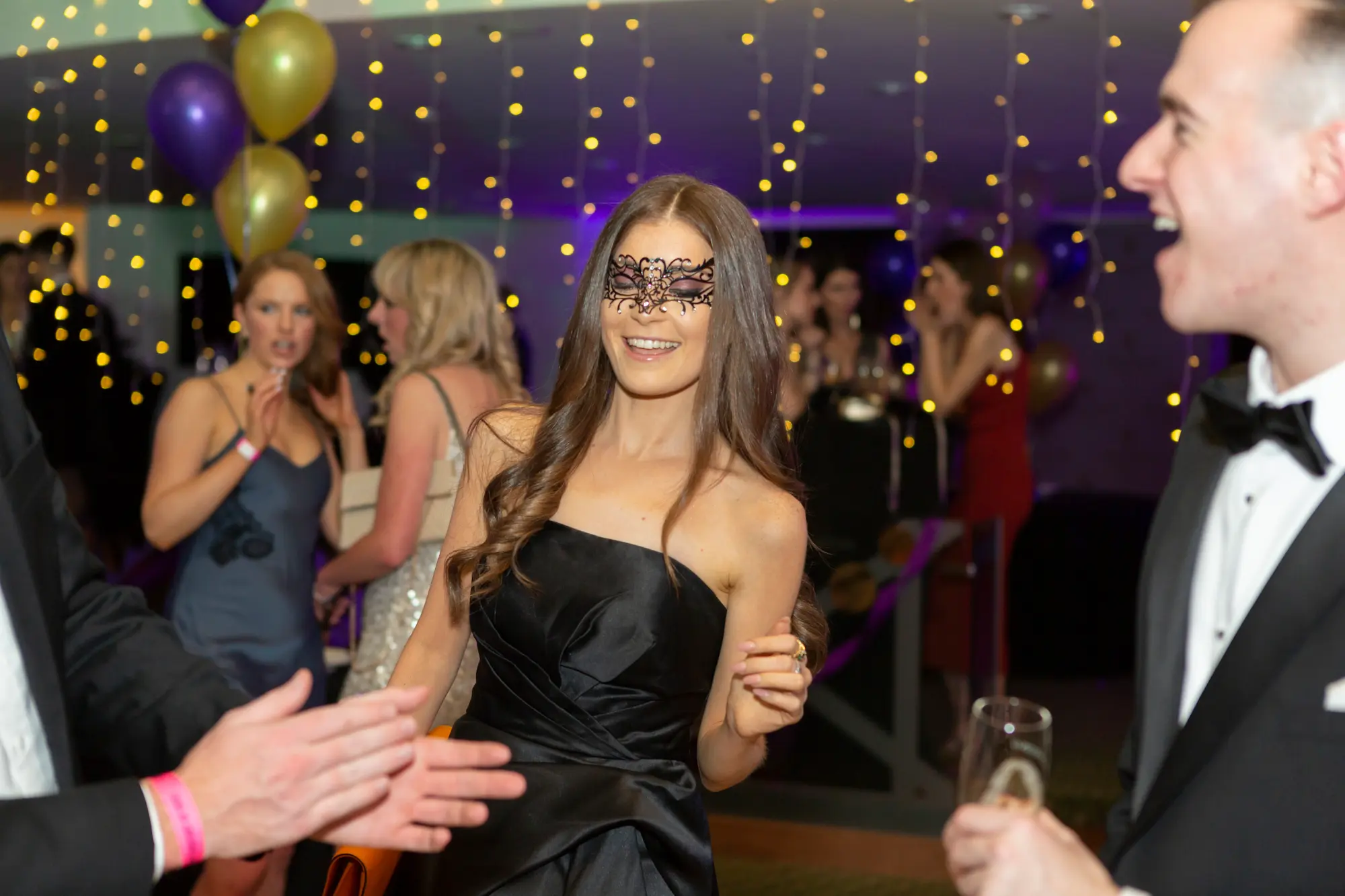 kooyong lawn tennis club masquerade ball black lace mask dance A lively and glamorous dance floor moment at the Kooyong Lawn Tennis Club End-of-Year Masquerade Ball, featuring a woman in a black strapless dress and intricate black lace eye mask smiling and interacting with guests. A man in a black tuxedo laughs while holding a champagne flute, and other formally dressed guests mingle nearby. The room is illuminated with vibrant purple lighting, warm fairy lights overhead, and clusters of gold and purple balloons, capturing the high-energy, elegant, and festive atmosphere of a sophisticated masquerade party.