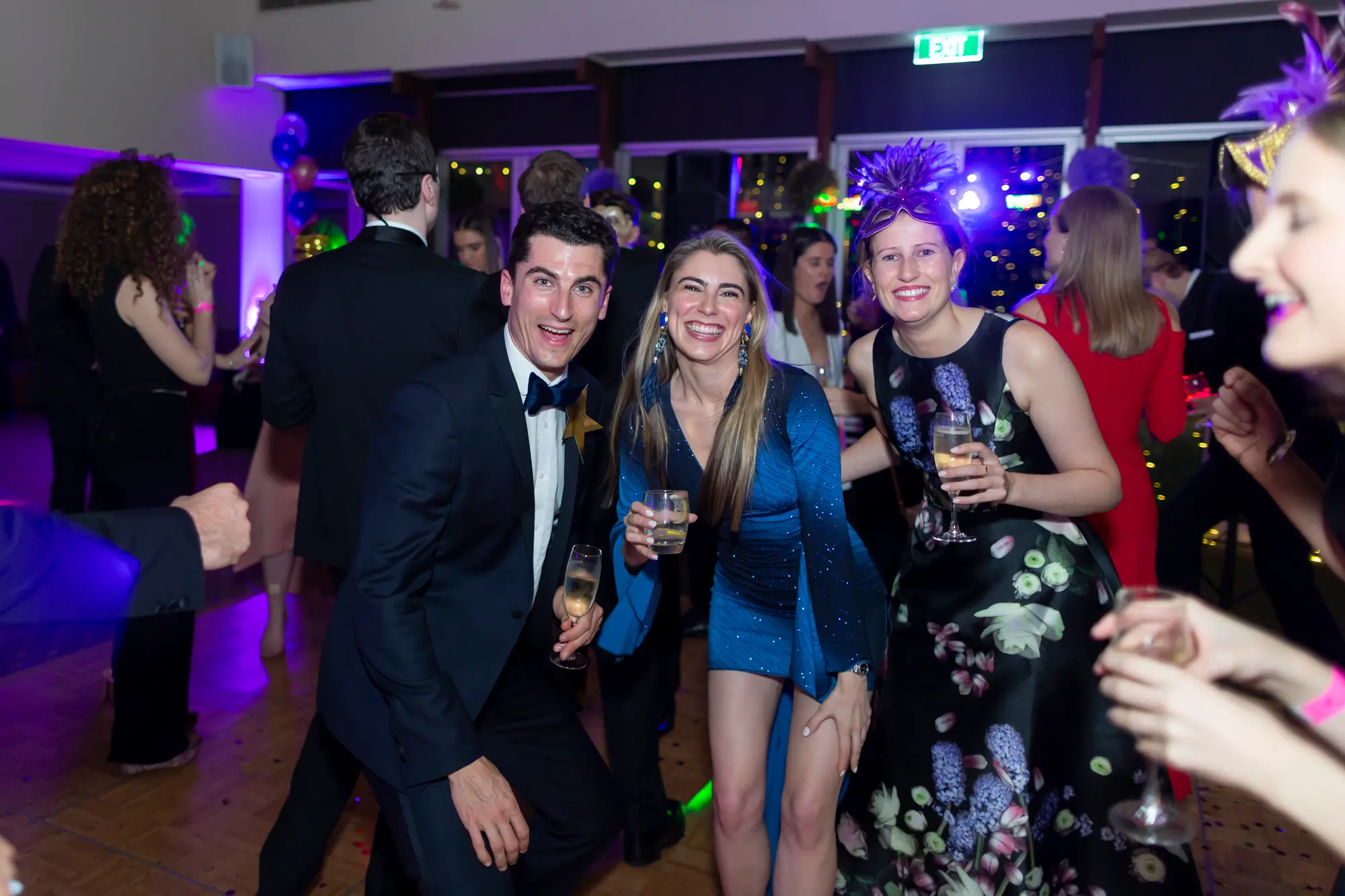 kooyong lawn tennis club masquerade ball blue tuxedo sequined mini dress dancefloor trio Three guests pose energetically on the dance floor at the Kooyong Lawn Tennis Club End-of-Year Masquerade Ball, featuring a man in a blue tuxedo crouching with champagne, a woman in a blue sequined mini dress playfully posing beside him, and a woman in a black floral dress holding a champagne flute. The lively indoor function room with dancing guests and colorful lighting creates a joyful black-tie party atmosphere.
