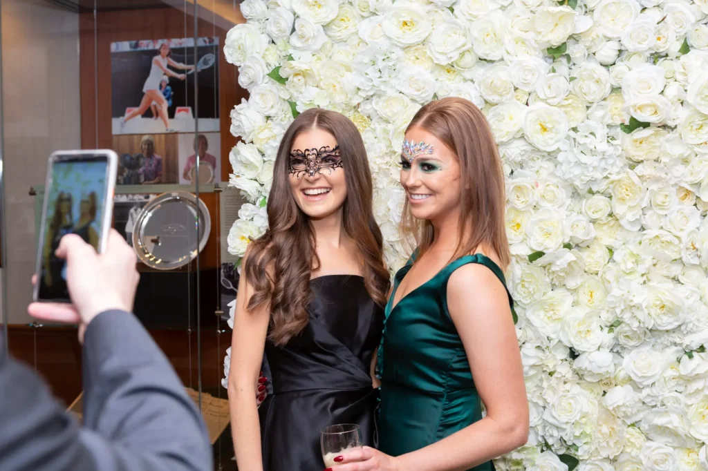 kooyong lawn tennis club masquerade ball candid guests floral wall photo moment A candid moment at the Kooyong Lawn Tennis Club End-of-Year Masquerade Ball shows two elegantly dressed guests posing and smiling in front of a striking white rose floral wall, both holding glasses of red wine and wearing ornate masquerade masks. Reflections of club memorabilia, including tennis photography and a silver trophy, appear through the glass nearby, grounding the scene in Kooyong’s rich sporting heritage. In the foreground, a guest’s smartphone captures the moment in real time, highlighting the lively, joyful atmosphere and the spontaneous social interactions that define this sophisticated end-of-year celebration.