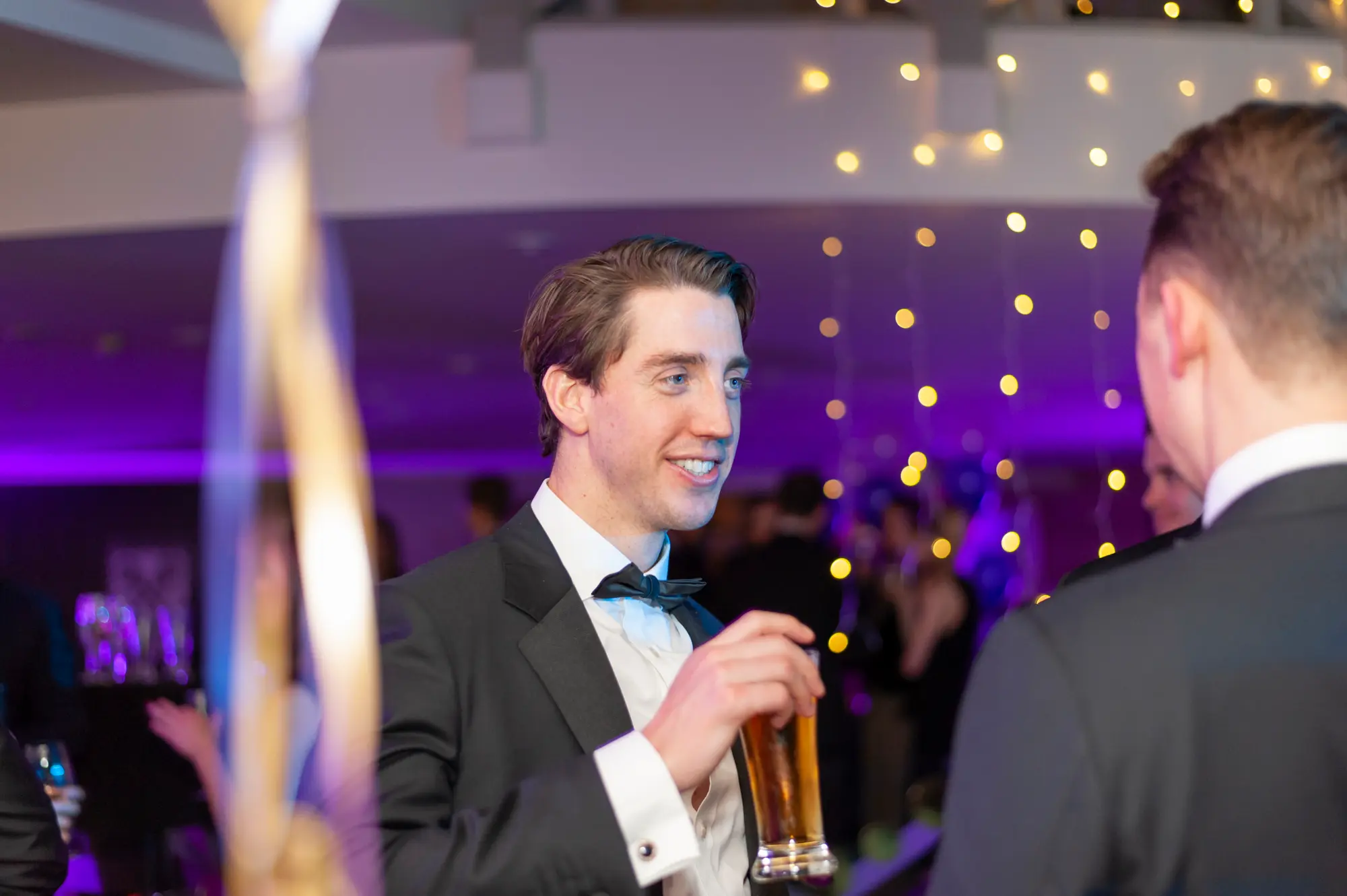 kooyong lawn tennis club masquerade ball candid two men conversation drinks A candid moment at the Kooyong Lawn Tennis Club End-of-Year Masquerade Ball captures two guests in black-tie attire sharing a relaxed conversation while enjoying drinks. Dressed in classic tuxedos with black bow ties and masquerade eye masks, one man holds a glass of beer as he turns toward his companion mid-conversation. Warm string lights glow overhead, with purple uplighting, framed artwork, and hints of the dance floor and entertainment setup in the background, reflecting the refined yet lively atmosphere of this elegant masquerade celebration.
