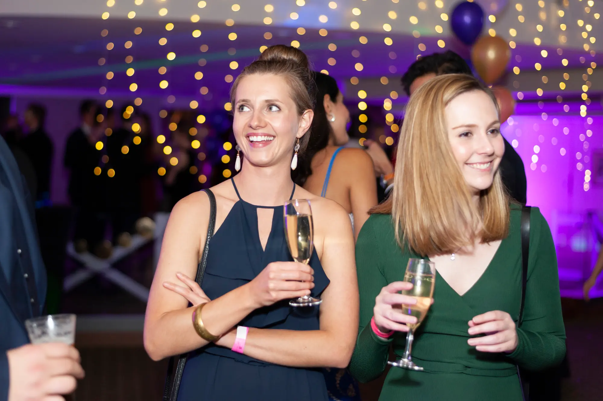 kooyong lawn tennis club masquerade ball candid two women champagne conversation A candid moment at the Kooyong Lawn Tennis Club End-of-Year Masquerade Ball shows two guests sharing a relaxed conversation while holding glasses of champagne. One woman wears a navy blue evening dress with statement drop earrings, while the other is dressed in an elegant emerald green V-neck gown with a shoulder bag. Warm string lights glow overhead, with purple uplighting, balloons, and mingling guests softly blurred in the background, capturing the lively yet refined atmosphere of this black-tie masquerade celebration in motion.