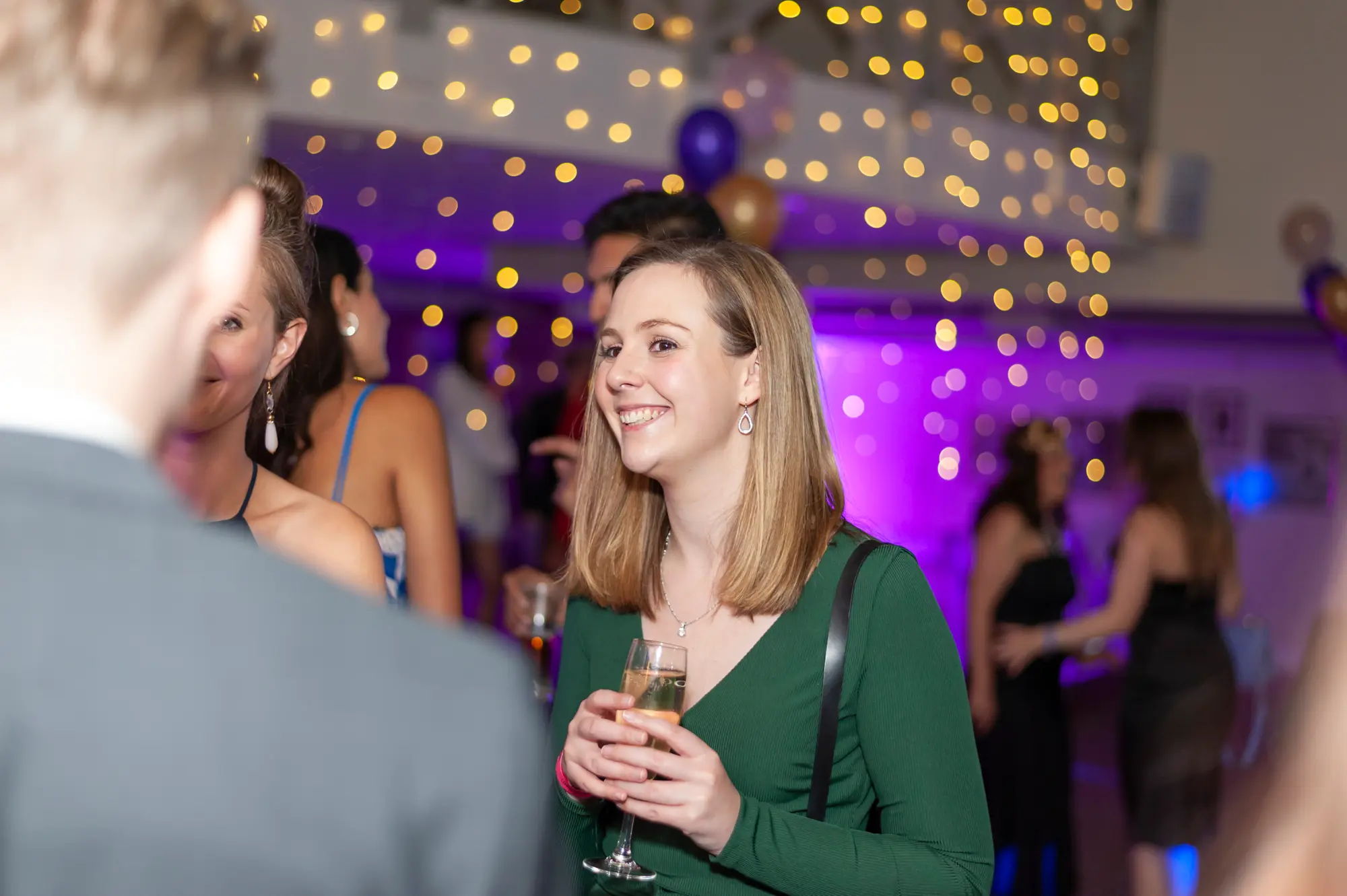 kooyong lawn tennis club masquerade ball candid woman green dress champagne A candid portrait of a guest enjoying the Kooyong Lawn Tennis Club End-of-Year Masquerade Ball, captured as she smiles warmly while holding a glass of champagne and chatting with another attendee. Dressed in an elegant emerald green evening gown with drop earrings, she stands beneath glowing string lights and soft purple uplighting, with balloons and mingling guests blurred in the background. The relaxed pose, ambient lighting, and lively social setting reflect the refined, celebratory atmosphere of the black-tie masquerade ball in motion.