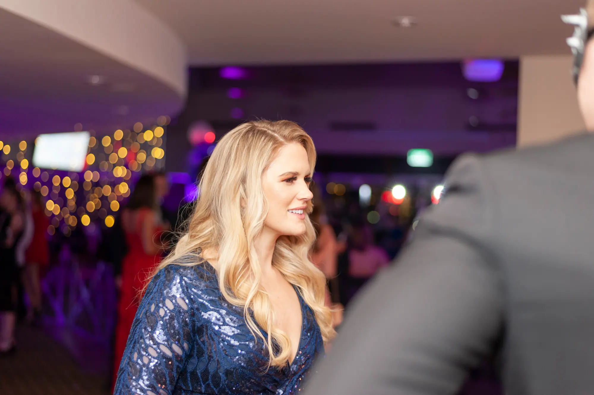 kooyong lawn tennis club masquerade ball candid woman sequined dress conversation A candid portrait from the Kooyong Lawn Tennis Club End-of-Year Masquerade Ball shows a guest in a navy blue sequined evening dress smiling warmly as she turns toward a conversation off-camera. Soft golden string lights create a glowing bokeh overhead, while purple uplighting washes the clubhouse walls and other formally dressed guests mingle in the background. Captured mid-interaction, this moment reflects the elegant, social energy and refined atmosphere of the black-tie masquerade celebration.