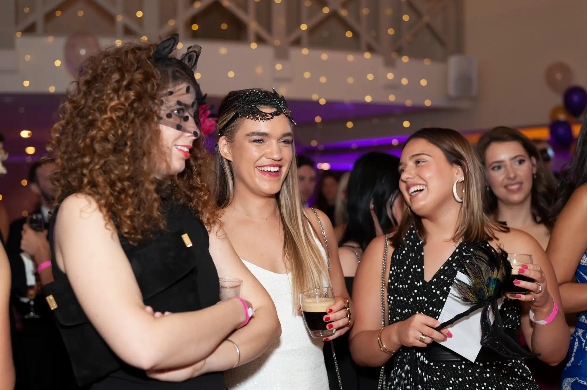 kooyong lawn tennis club masquerade ball candid women posing feather mask Three non-celebrity women pose together during candid moments at the Kooyong Lawn Tennis Club End-of-Year Masquerade Ball, standing confidently beneath high open-trussed ceilings with soft purple ambient lighting. Dressed in elegant formal evening wear, the women wear masquerade eye masks, including black lace masks and a peacock-feathered mask accessory, while holding drinks and smiling naturally for the camera. The relaxed poses, modern pendant lighting, and mingling guests in the background capture the sophisticated, social atmosphere of the black-tie masquerade celebration in motion.