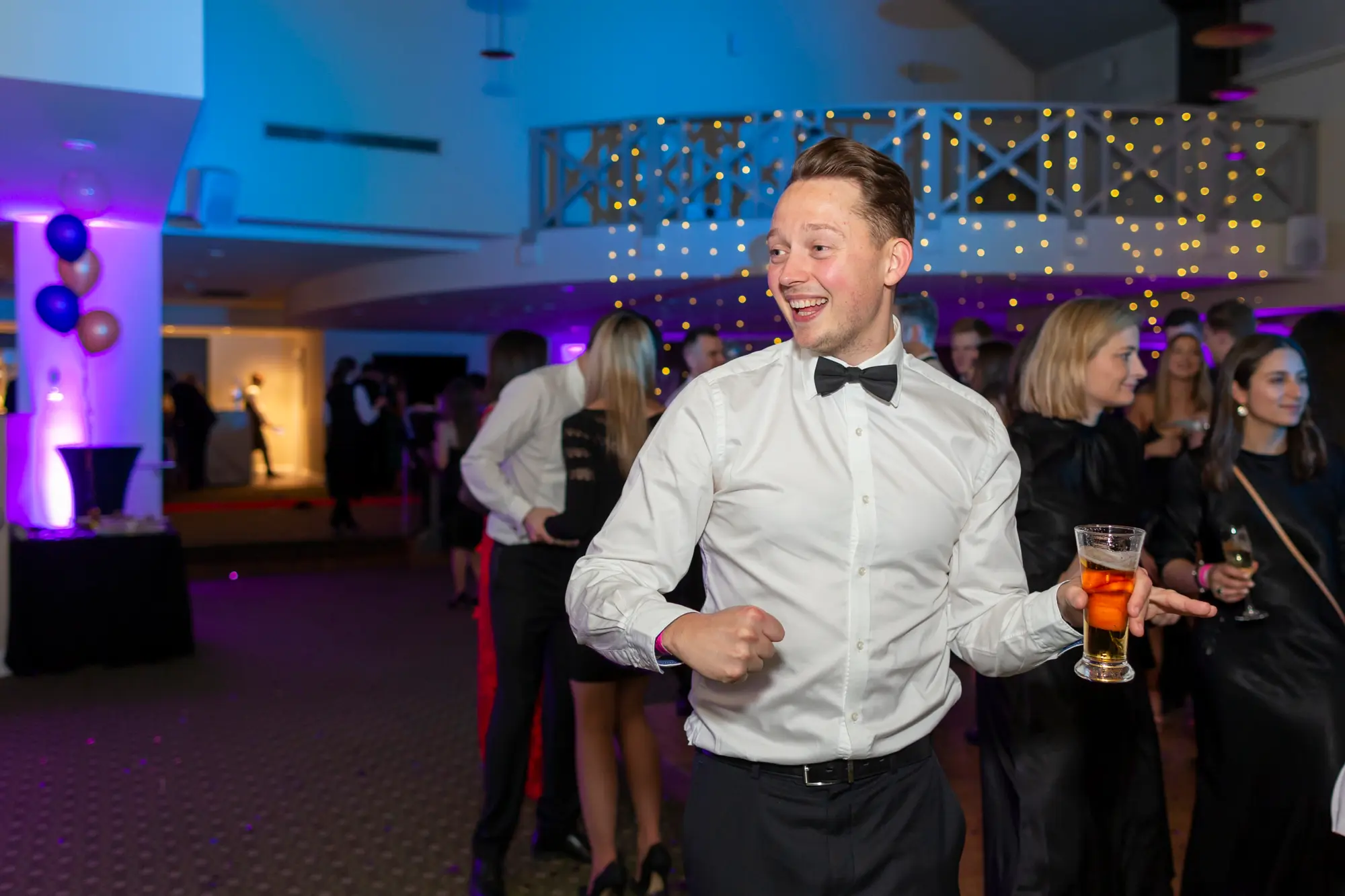 kooyong lawn tennis club masquerade ball dance floor celebration smile man Energetic guests on the dance floor at Kooyong Lawn Tennis Club Masquerade Ball, including a man in a white shirt and black bow tie dancing with a glass of amber drink, a woman in a black lace-trimmed slip dress laughing with a champagne flute, and other formally dressed guests, with purple and blue lighting, raised hands, and festive decorations