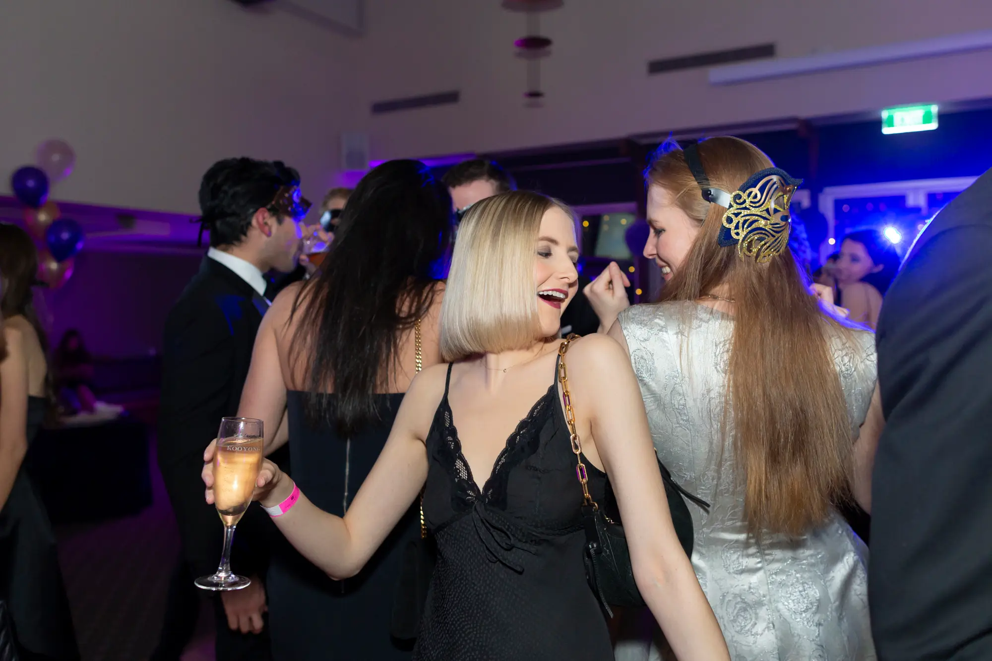 kooyong lawn tennis club masquerade ball dance floor celebration Guests dancing energetically on the dance floor at Kooyong Lawn Tennis Club Masquerade Ball with vibrant blue lighting and string lights, a blonde woman in a black lace-trim slip dress holding a champagne flute, a red-haired woman in an elaborate gold and blue masquerade mask smiling, and other formally dressed guests mingling and dancing