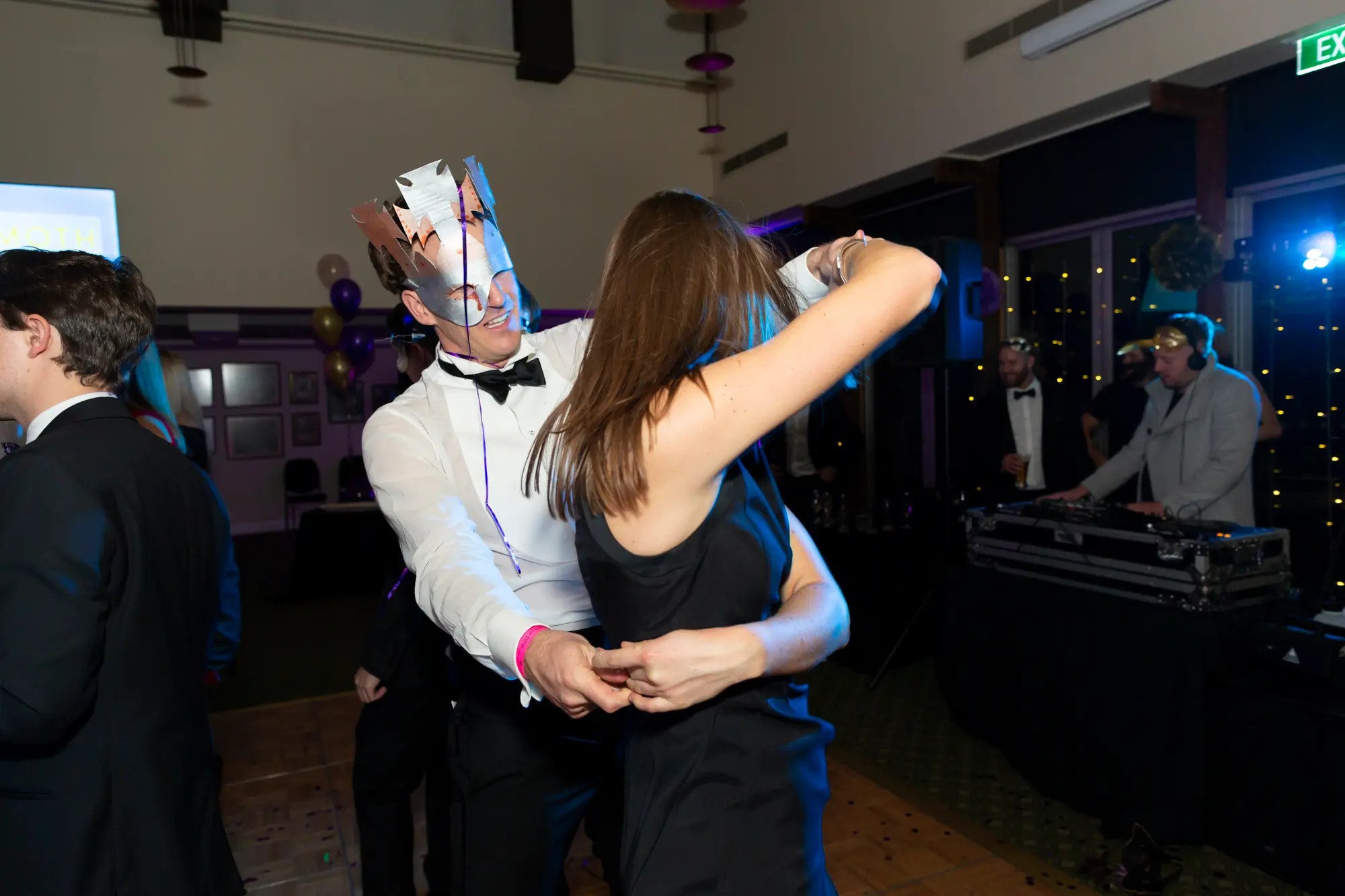 kooyong lawn tennis club masquerade ball dance floor couple dancing Man in a white dress shirt, black bow tie, and festive paper crown dancing closely with a woman in a black sleeveless cocktail dress on the wooden dance floor at Kooyong Lawn Tennis Club Masquerade Ball, with a DJ in the background and other formally dressed guests enjoying the celebration