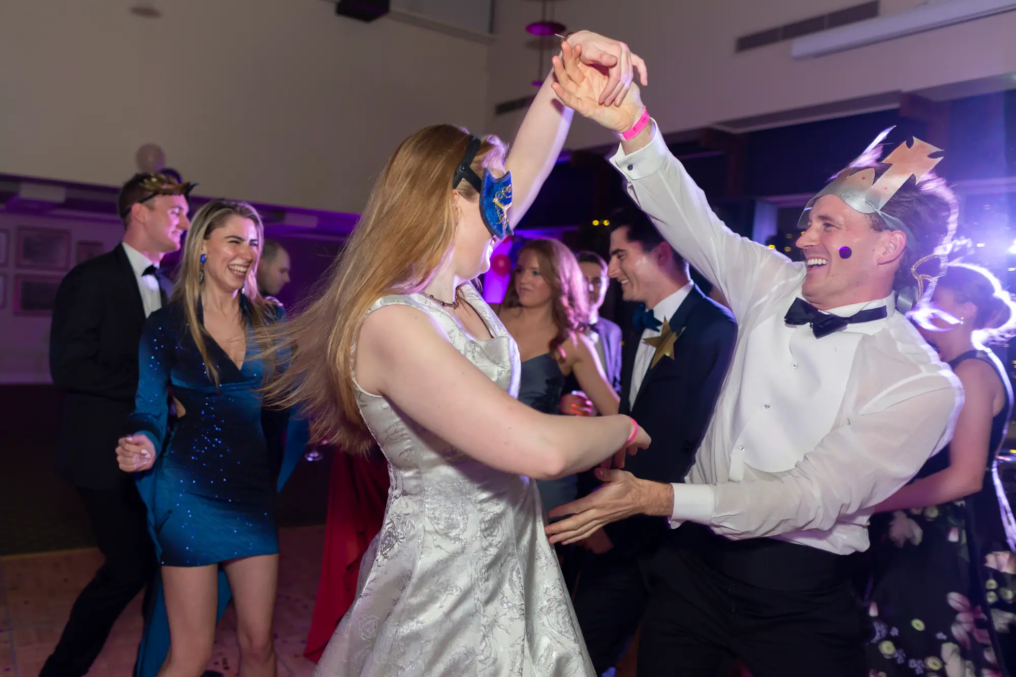 kooyong lawn tennis club masquerade ball dance floor couple spin Man wearing a white dress shirt, black bow tie, and colorful paper crown dancing with a woman in a white patterned dress and blue eye mask on the wooden dance floor at Kooyong Lawn Tennis Club Masquerade Ball, with other formally dressed guests, purple stage lighting, and a lively party atmosphere in the background