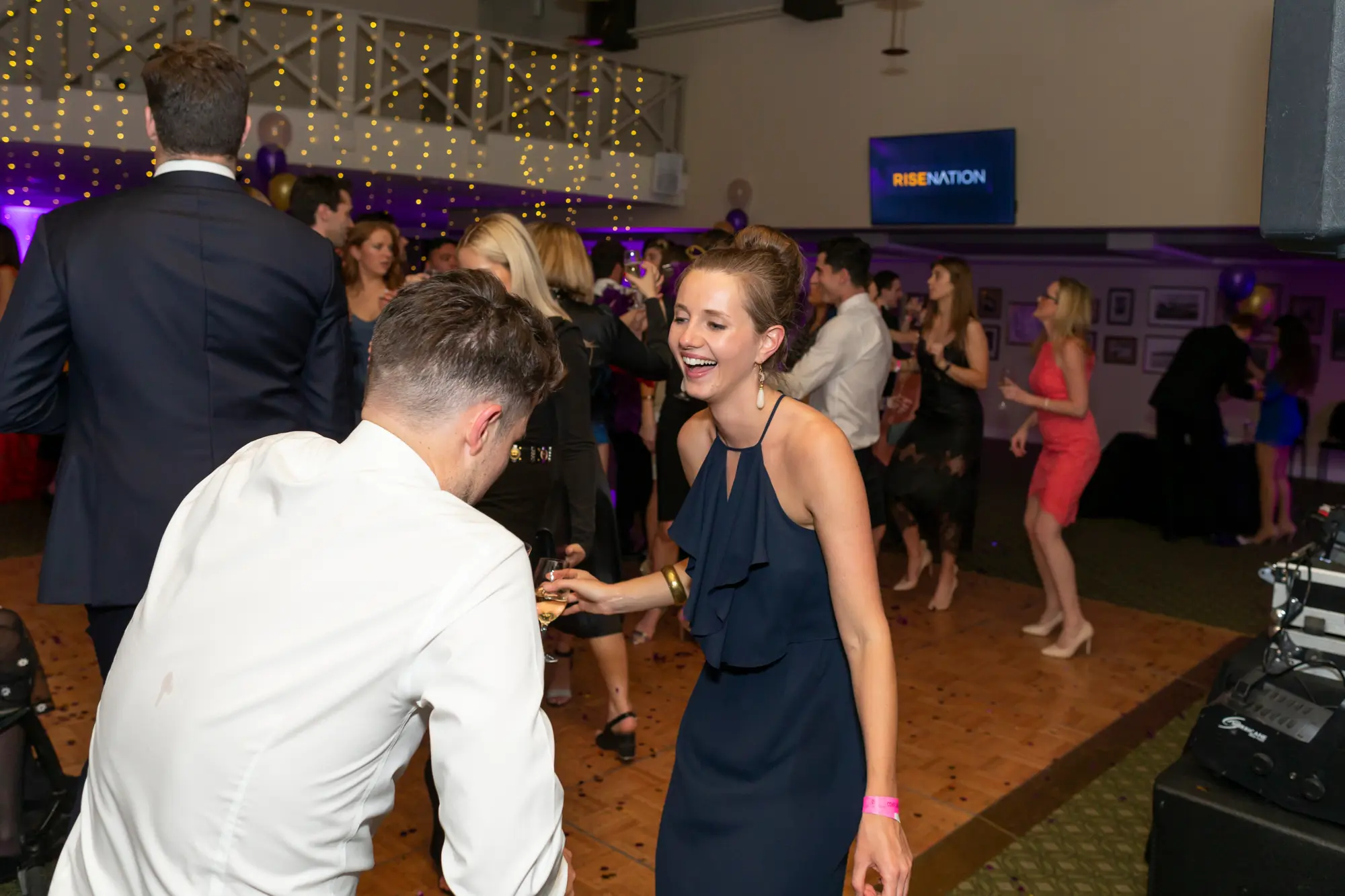 kooyong lawn tennis club masquerade ball dance floor dancing couple joyful A lively and energetic moment on the dance floor at the Kooyong Lawn Tennis Club End-of-Year Masquerade Ball, featuring a man in a white shirt and black trousers dancing with a laughing woman in a navy blue ruffled halter-neck dress holding a champagne flute. Guests in tuxedos and cocktail dresses mingle under vibrant purple lighting, with fairy lights draped across the mezzanine and clusters of purple and gold balloons adding a festive touch. The scene captures joyful celebration, high-energy dancing, and the glamorous, sophisticated atmosphere of a black-tie masquerade party.