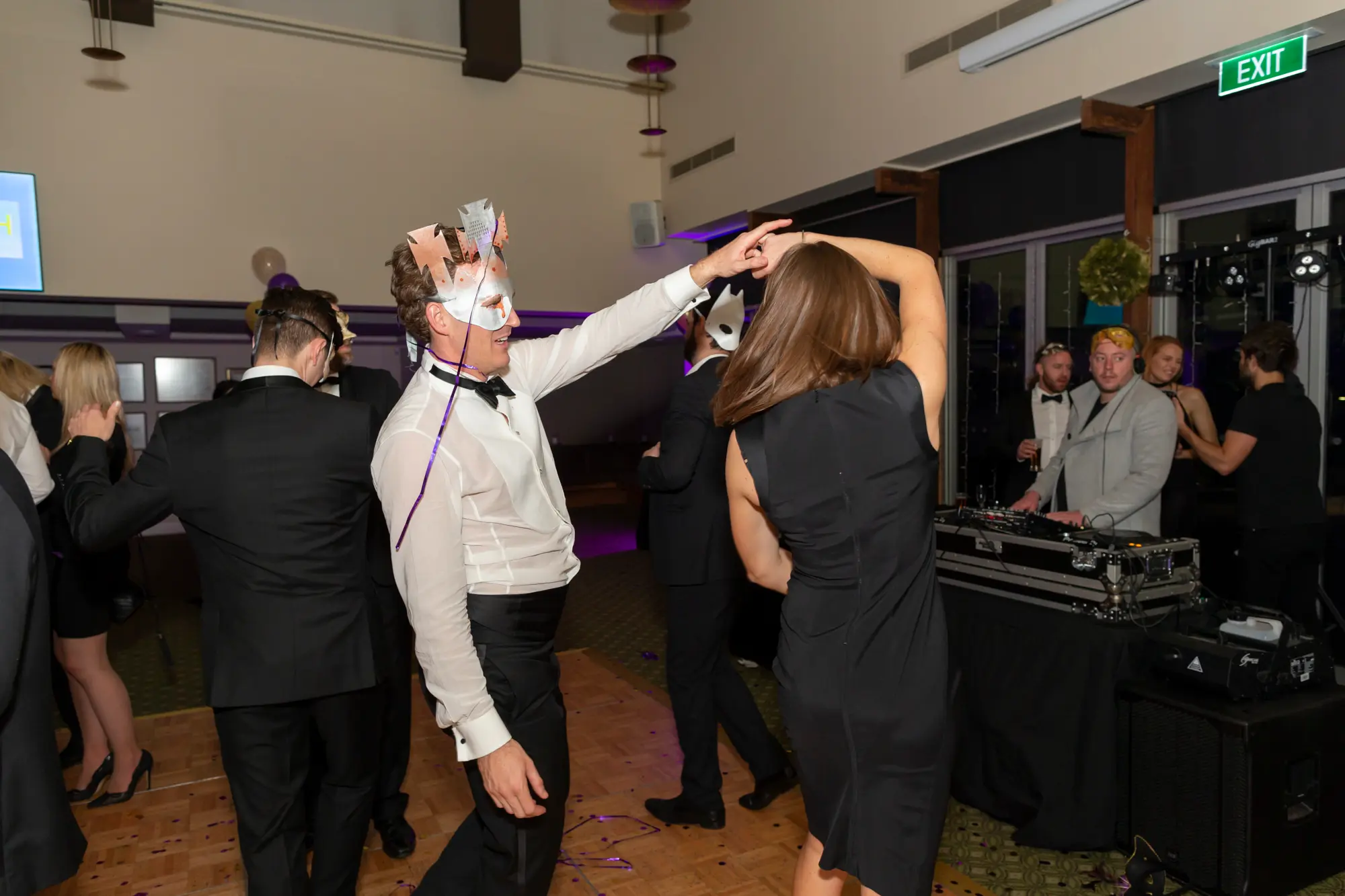 kooyong lawn tennis club masquerade ball dance floor dancing couple Man in a white dress shirt, black bow tie, and white masquerade mask dancing with a woman in a black sleeveless dress on the wooden dance floor at Kooyong Lawn Tennis Club Masquerade Ball, with a DJ booth and other formally dressed guests in the background, surrounded by confetti and ambient lighting