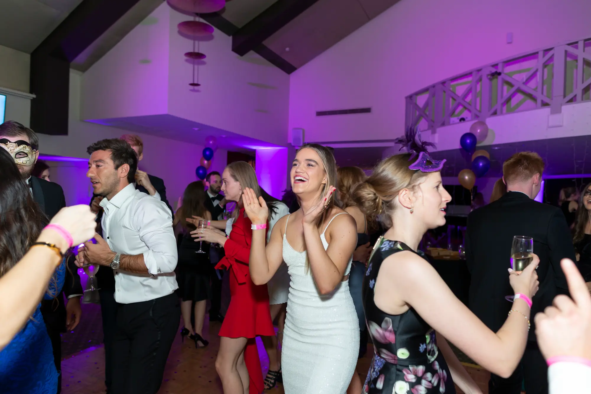 kooyong lawn tennis club masquerade ball dance floor group celebration Group of guests dancing and celebrating on the wooden dance floor at Kooyong Lawn Tennis Club Masquerade Ball, including a woman in a white dress laughing, a woman in a red dress holding a glass, and men in tuxedos, with purple ambient lighting, fairy lights, and scattered balloons creating a lively party atmosphere