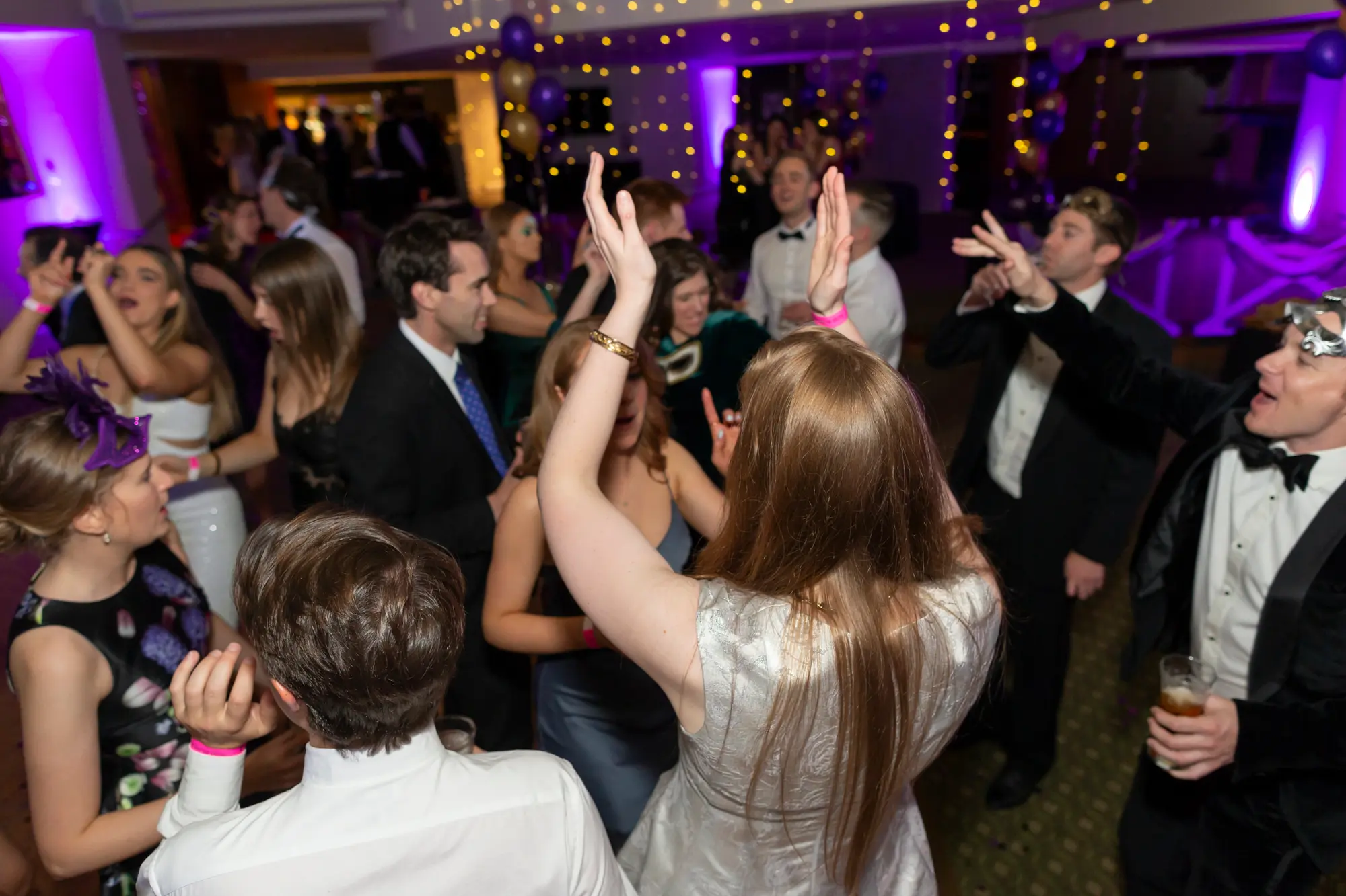 kooyong lawn tennis club masquerade ball dance floor party energy Guests dancing energetically on the wooden dance floor at Kooyong Lawn Tennis Club Masquerade Ball, including a woman in a blue lace dress cheering, a man in a black tuxedo with a silver masquerade mask holding a drink, and a woman in a silver sequined dress, with purple stage lighting, fairy lights, and balloons creating a lively festive atmosphere.