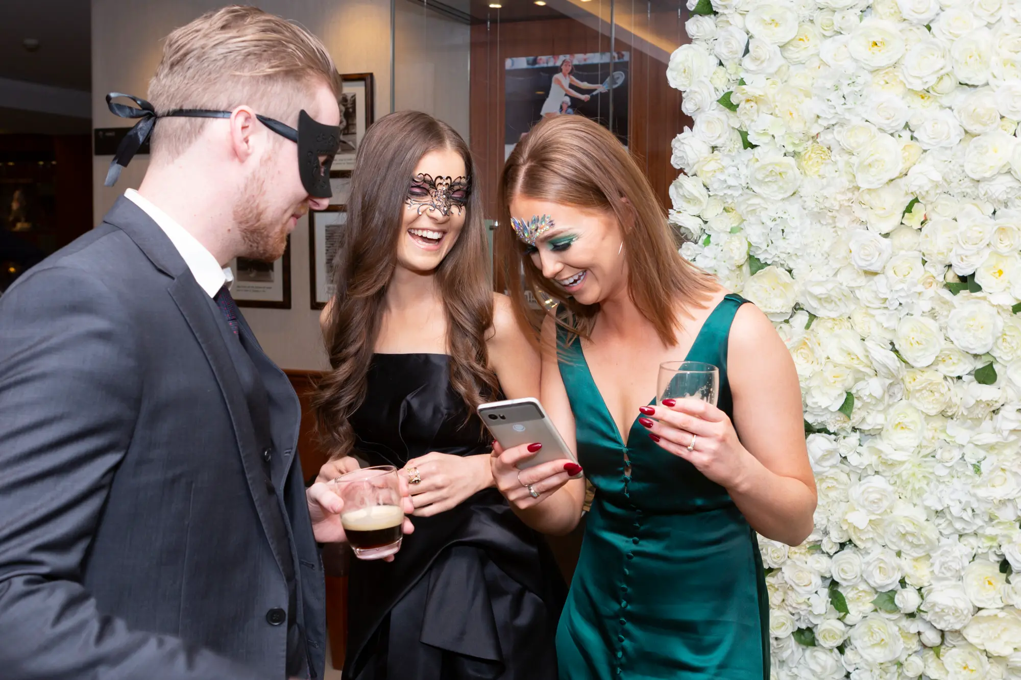 kooyong lawn tennis club masquerade ball floral wall candid guest portrait A candid and joyful moment at the Kooyong Lawn Tennis Club End-of-Year Masquerade Ball in Toorak, showing two elegantly dressed women posing in front of a floor-to-ceiling white rose floral wall while holding red drinks and wearing black masquerade masks. A third guest in a suit and mask is partially visible beside them, adding to the lively social atmosphere. In the foreground, a guest’s smartphone is seen capturing the photo, with reflections of tennis memorabilia and trophies behind glass reinforcing the prestigious clubhouse setting and the festive elegance of the evening.