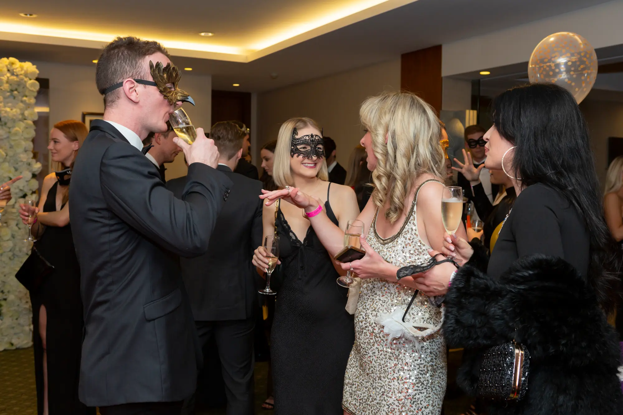 kooyong lawn tennis club masquerade ball floral wall social candid interaction A candid social moment at the Kooyong Lawn Tennis Club End-of-Year Masquerade Ball in Toorak, showing guests gathered in front of a white rose floral wall, enjoying drinks and conversation in elegant evening wear and masquerade masks. Two women in black and gold dresses smile while chatting, as a man in a suit and gold mask sips champagne nearby, with reflective glass walls and tennis memorabilia subtly framing the lively, sophisticated atmosphere of the celebration.