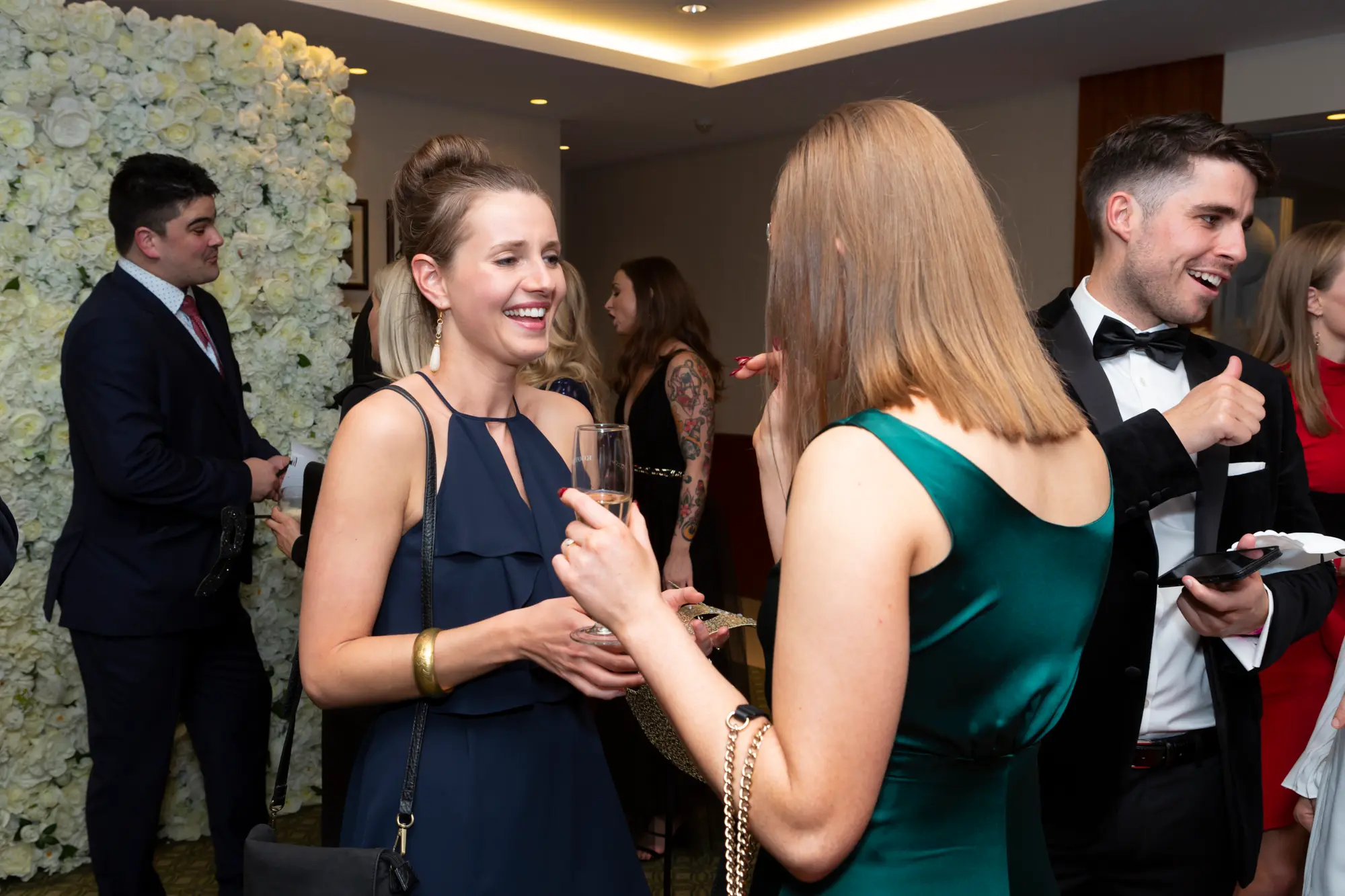 kooyong lawn tennis club masquerade ball floral wall three women black tie Three women in elegant evening gowns pose and smile in front of a white rose floral wall at the Kooyong Lawn Tennis Club End-of-Year Masquerade Ball in Toorak, capturing a festive black-tie atmosphere with warm ambient lighting, reflective glass panels, tennis memorabilia, and guests enjoying drinks and lively social interaction.