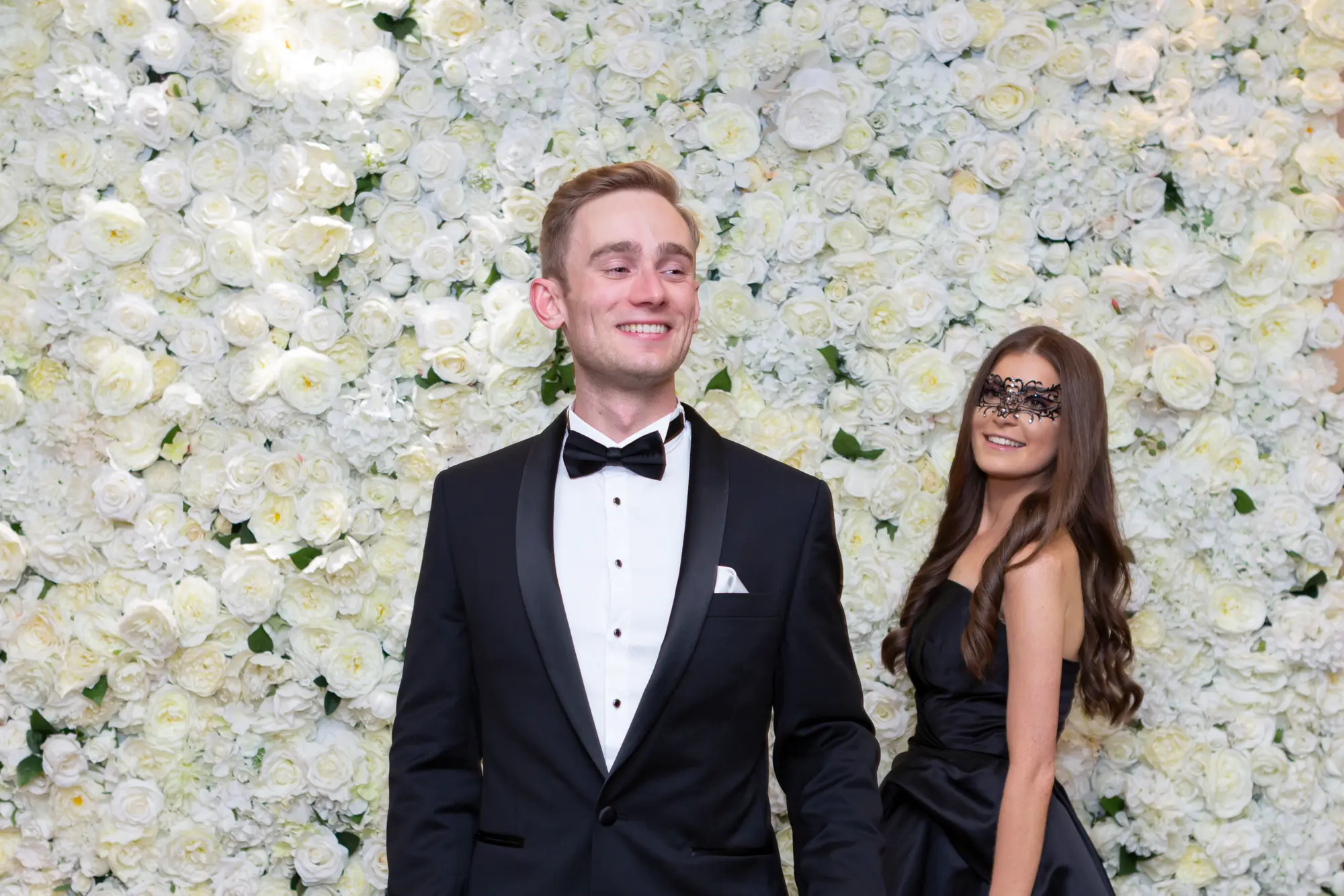kooyong lawn tennis club masquerade ball flower wall elegant couple portrait An elegant couple poses in front of a white flower wall at the Kooyong Lawn Tennis Club End-of-Year Masquerade Ball. The man wears a classic black tuxedo and smiles slightly off-camera, while the woman in a strapless black dress and black lace masquerade mask looks up at him warmly. The floor-to-ceiling floral backdrop creates a bright, romantic contrast to their dark formal attire, capturing a joyful and sophisticated moment from the glamorous celebration.