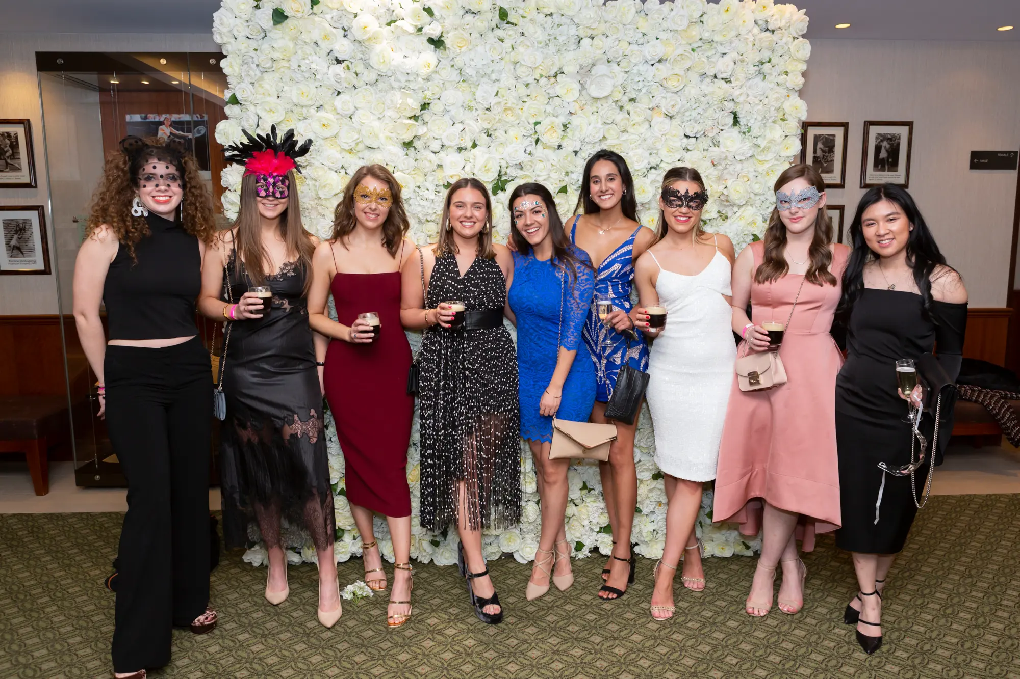 kooyong lawn tennis club masquerade ball group portrait eight women floral wall Eight women pose together at the Kooyong Lawn Tennis Club End-of-Year Masquerade Ball in Toorak, gathered in front of an elegant floor-to-ceiling white rose floral wall. Dressed in black-tie evening gowns in black, red, green, blue, pink, and gold sequined styles, they smile brightly while holding drinks and wearing ornate masquerade masks. Warm ambient lighting, reflective interior surfaces, and subtle tennis club memorabilia create a sophisticated and celebratory atmosphere, capturing the social energy of this iconic Melbourne masquerade event.