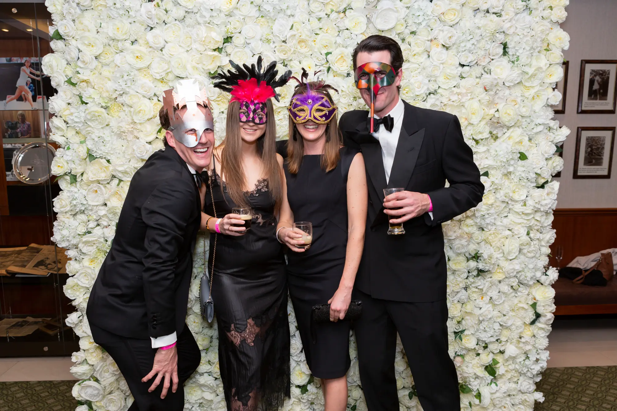 kooyong lawn tennis club masquerade ball group portrait four guests floral wall Four guests pose together for a lively group portrait at the Kooyong Lawn Tennis Club End-of-Year Masquerade Ball in Toorak, smiling in front of a dramatic floor-to-ceiling white rose floral wall. Dressed in elegant black-tie attire, including black dresses, a blue-grey evening gown, and tuxedos with gold, silver, and black masquerade masks, the group shares a playful moment as one man leans forward in a relaxed pose. Warm ambient lighting, reflective interior details, and subtle tennis club memorabilia create a refined yet celebratory atmosphere at this prestigious masquerade event.