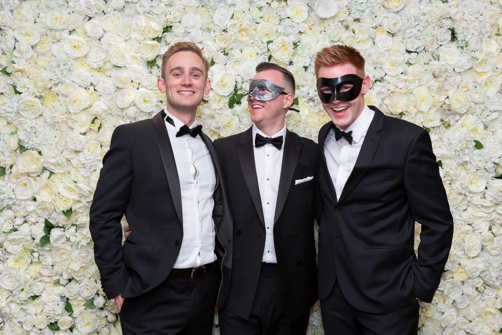 kooyong lawn tennis club masquerade ball group portrait men floral wall Three men pose together in black tuxedos during the Kooyong Lawn Tennis Club End-of-Year Masquerade Ball in Toorak, standing in front of a luxurious white rose floral wall. One guest playfully holds a silver masquerade mask to his face while the others smile with relaxed confidence, capturing the refined yet celebratory spirit of the evening. Warm ambient lighting, reflective walls, and tennis club memorabilia subtly frame the scene, highlighting the elegance, camaraderie, and black-tie glamour of this prestigious masquerade event at one of Melbourne’s most iconic venues.
