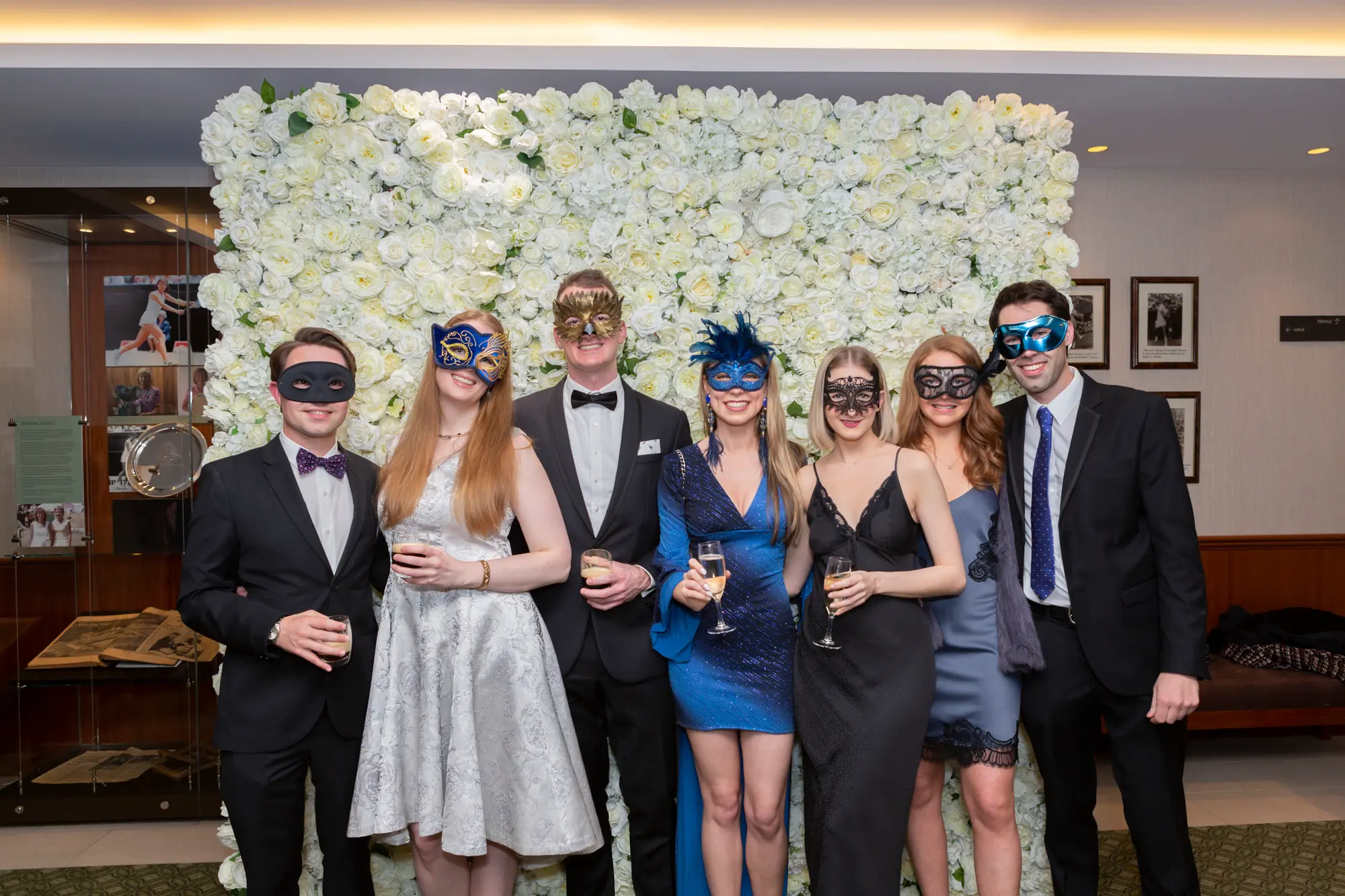 kooyong lawn tennis club masquerade ball group portrait seven guests floral wall Seven guests pose together at the Kooyong Lawn Tennis Club End-of-Year Masquerade Ball in Toorak, standing in front of a striking floor-to-ceiling white rose floral wall. Dressed in elegant black-tie evening wear in shades of black, navy blue, light blue-grey, and emerald green, they smile warmly while holding drinks and wearing decorative masquerade masks. Soft ambient lighting, reflective interior surfaces, and subtle tennis club memorabilia enhance the refined atmosphere, capturing the lively social spirit of this prestigious Melbourne masquerade event.