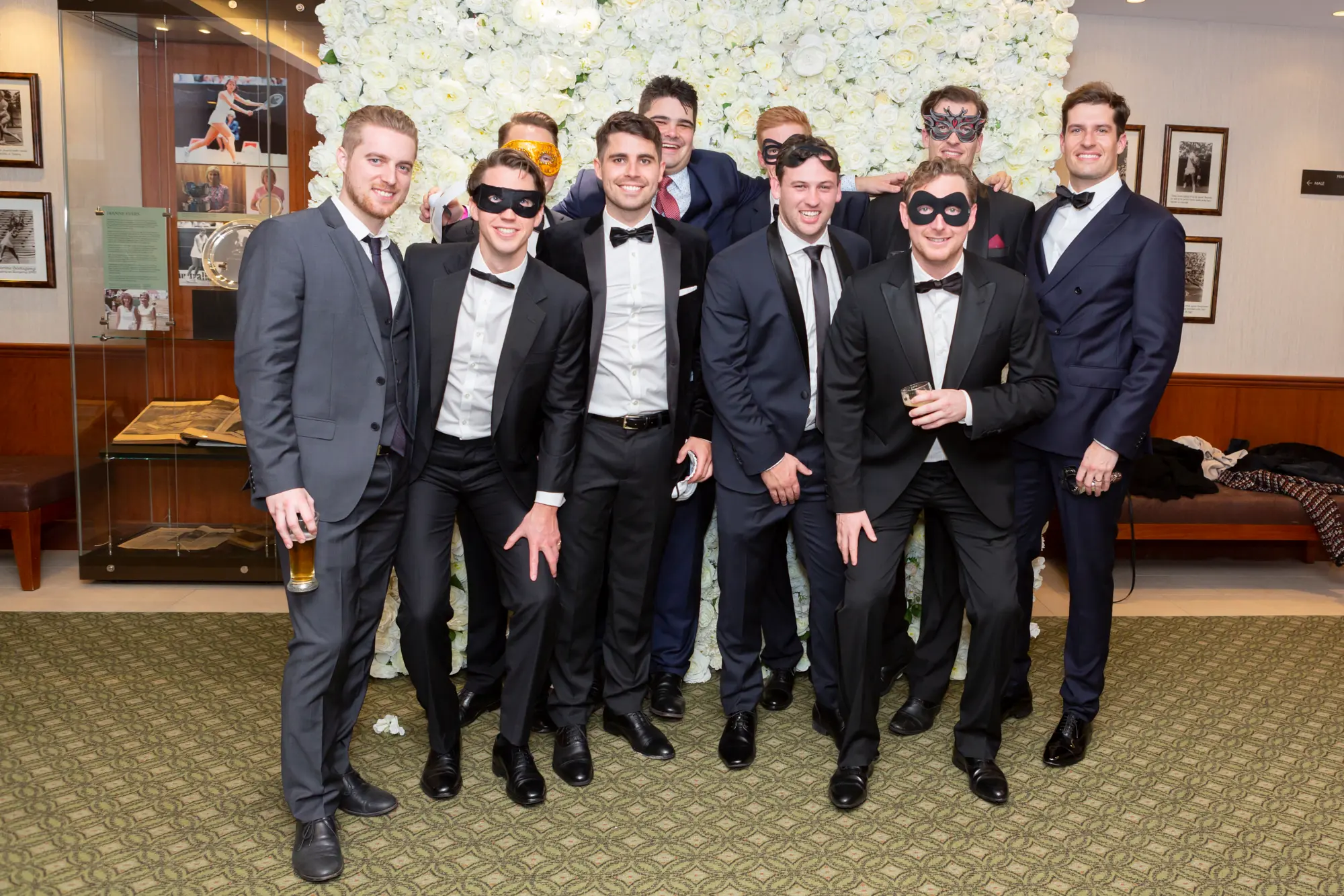 kooyong lawn tennis club masquerade ball group portrait ten men Ten male guests pose together for a relaxed group portrait at the Kooyong Lawn Tennis Club End-of-Year Masquerade Ball in Toorak, standing in front of an elegant floor-to-ceiling white rose floral wall. Dressed in black tuxedos and tailored grey suits with black bow ties, and wearing a mix of black, silver, and gold masquerade masks, the group smiles confidently, with one guest holding a drink and another standing casually with hands in his pockets. Warm ambient lighting, reflective interiors, and subtle tennis club memorabilia frame the sophisticated atmosphere of this prestigious end-of-year celebration.
