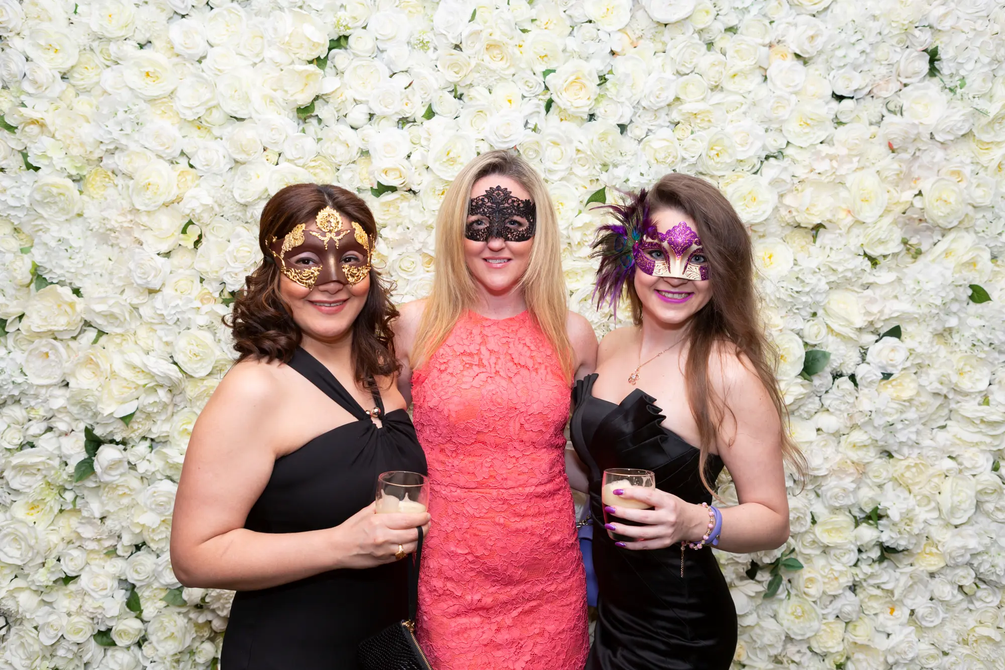kooyong lawn tennis club masquerade ball group portrait three women floral wall Three women pose together for a group portrait at the Kooyong Lawn Tennis Club End-of-Year Masquerade Ball in Toorak, smiling and enjoying drinks in front of an elegant floor-to-ceiling white rose floral wall. Dressed in formal evening gowns in black, coral-orange, and black with an emerald sheen, and wearing decorative masquerade masks, they embody the glamour of this prestigious black-tie celebration. Warm ambient lighting, reflective interior surfaces, and subtle tennis club memorabilia enhance the refined and festive atmosphere of the event.
